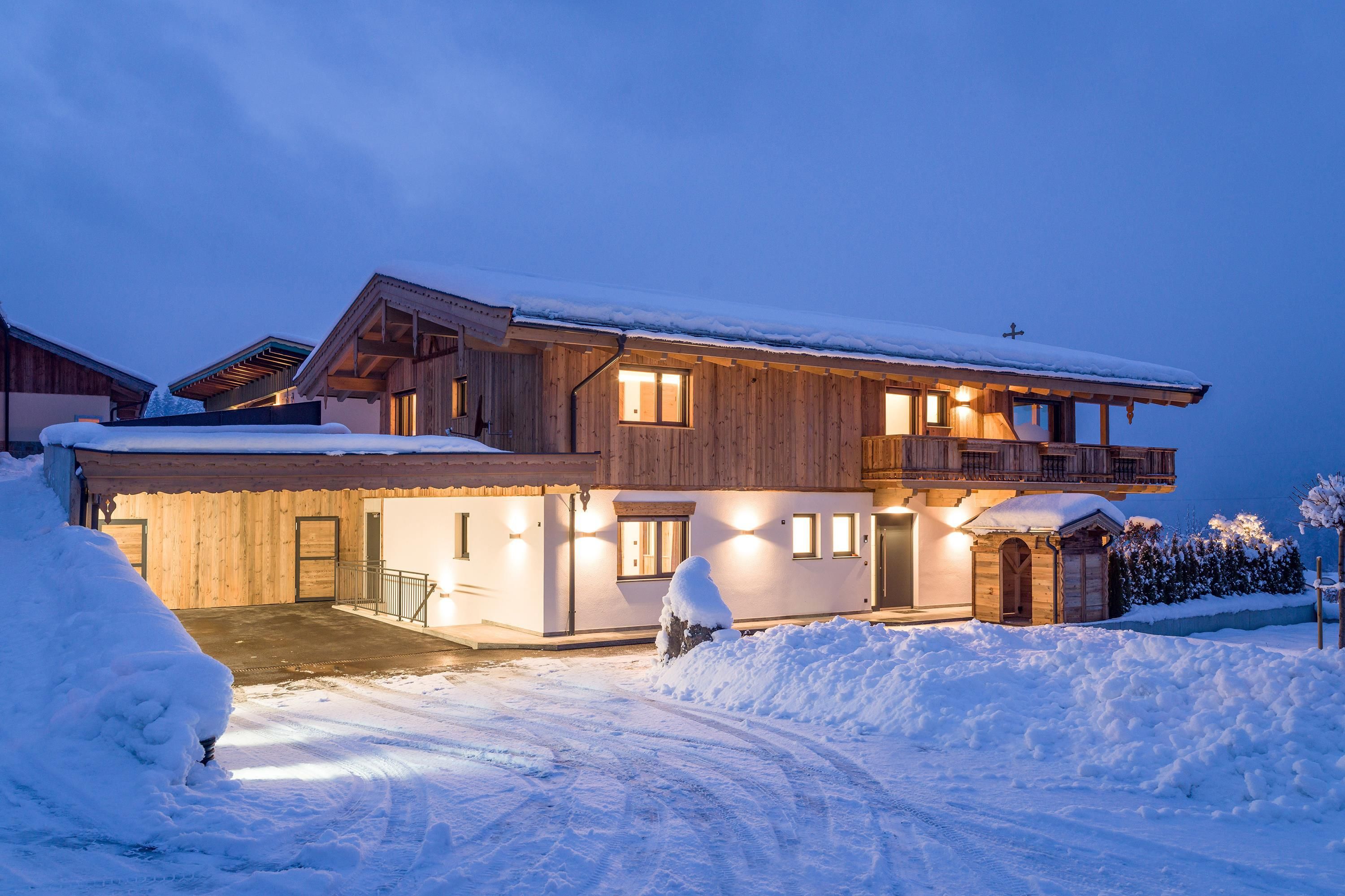 A modern wooden house in the snow, illuminated at dusk. Fresh, white snow surrounds it and the sky is gray-blue.