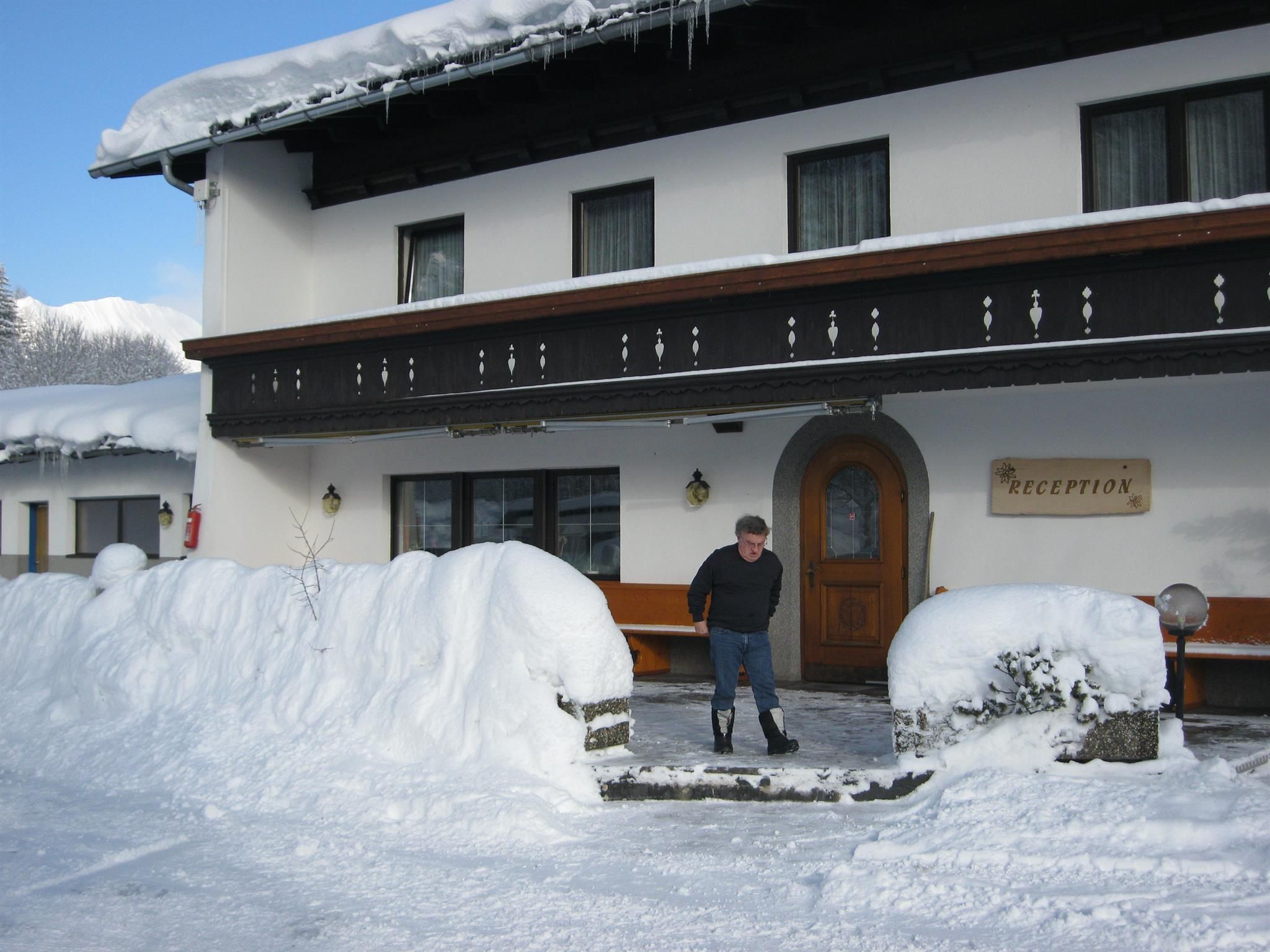 A cozy hotel in the snow with a man standing in front of the entrance door. The surroundings are wintry, with plenty of snow and a clear sky.