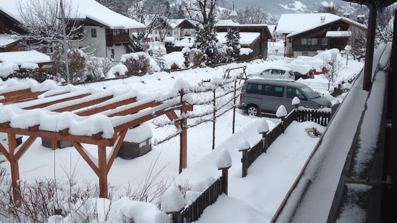 A snowy landscape with wooden structures and vehicles. In the background, cozy houses and snow-covered trees can be seen.