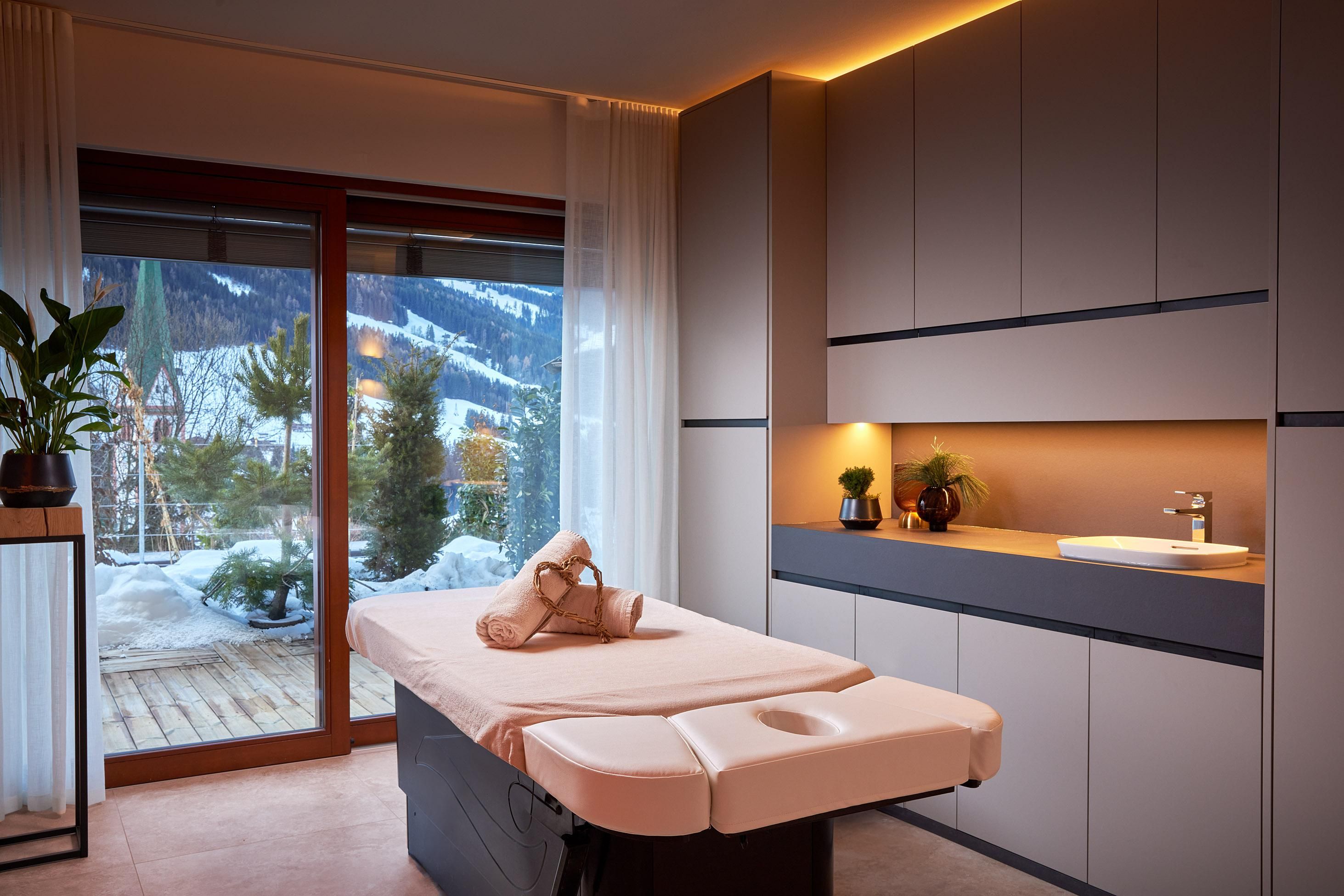 A modern spa room with a massage bed and soft lighting. Large windows offer a view of the snowy landscape.