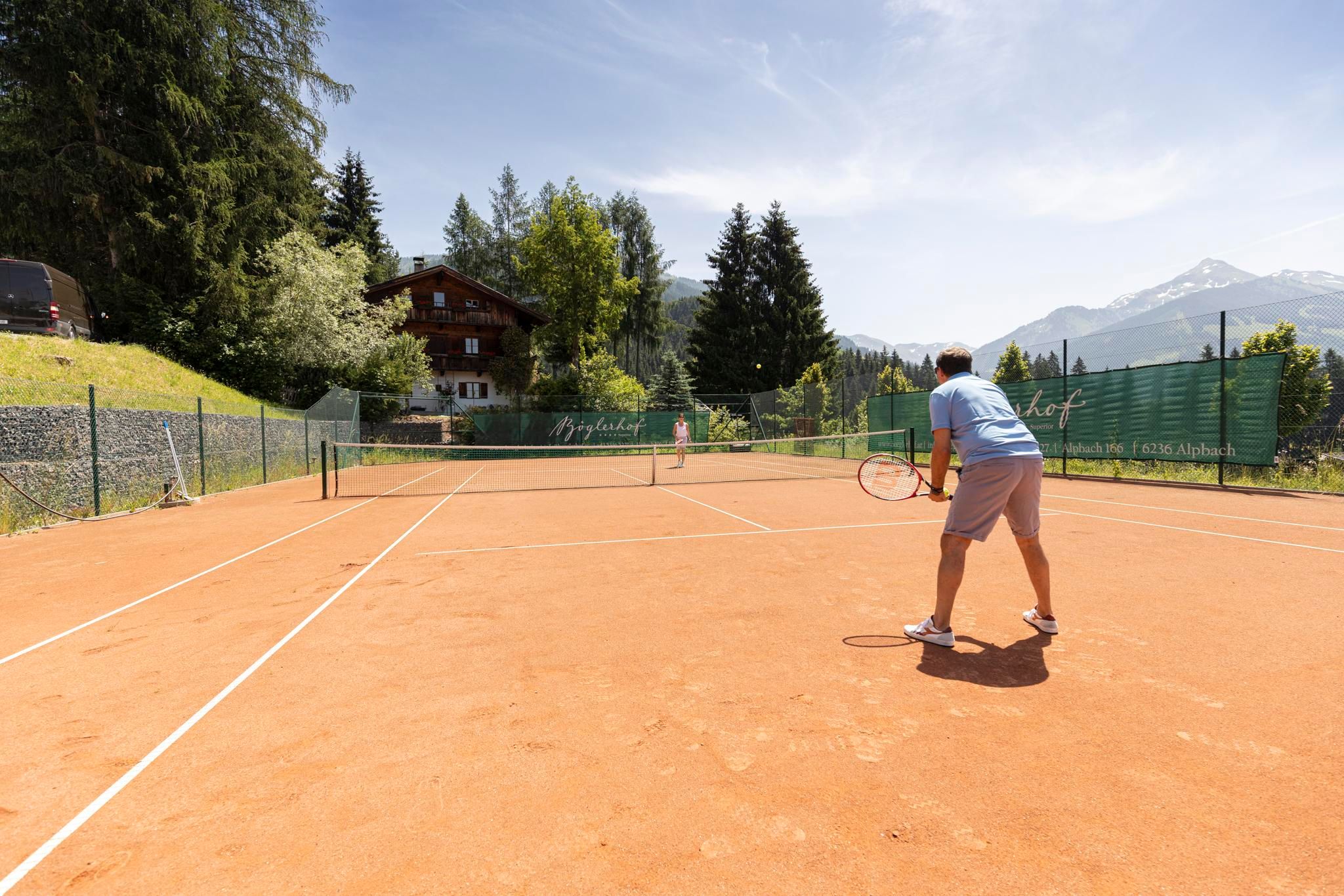A tennis court in the mountains with a player preparing for the serve. In the background, trees and a wooden house are visible.