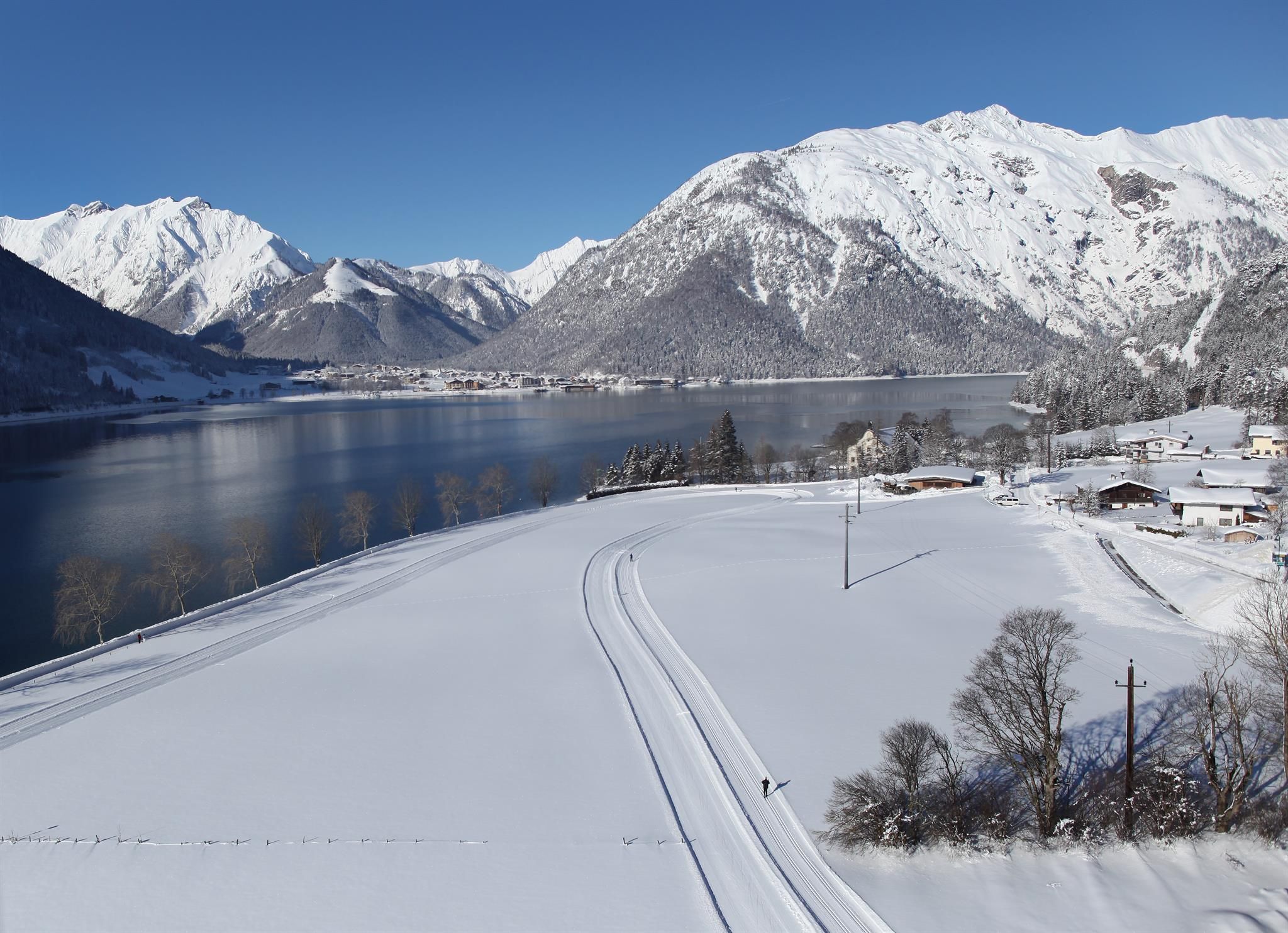 A snowy landscape with mountains in the background and a clear blue sky. A lake reflects the surroundings and creates a tranquil atmosphere.