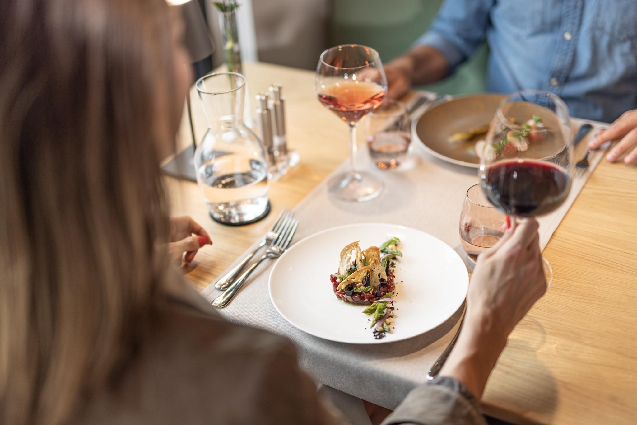An elegant restaurant with a beautifully arranged plate and glasses of red and rosé wine. In the background, a second person can be seen, also enjoying a meal.