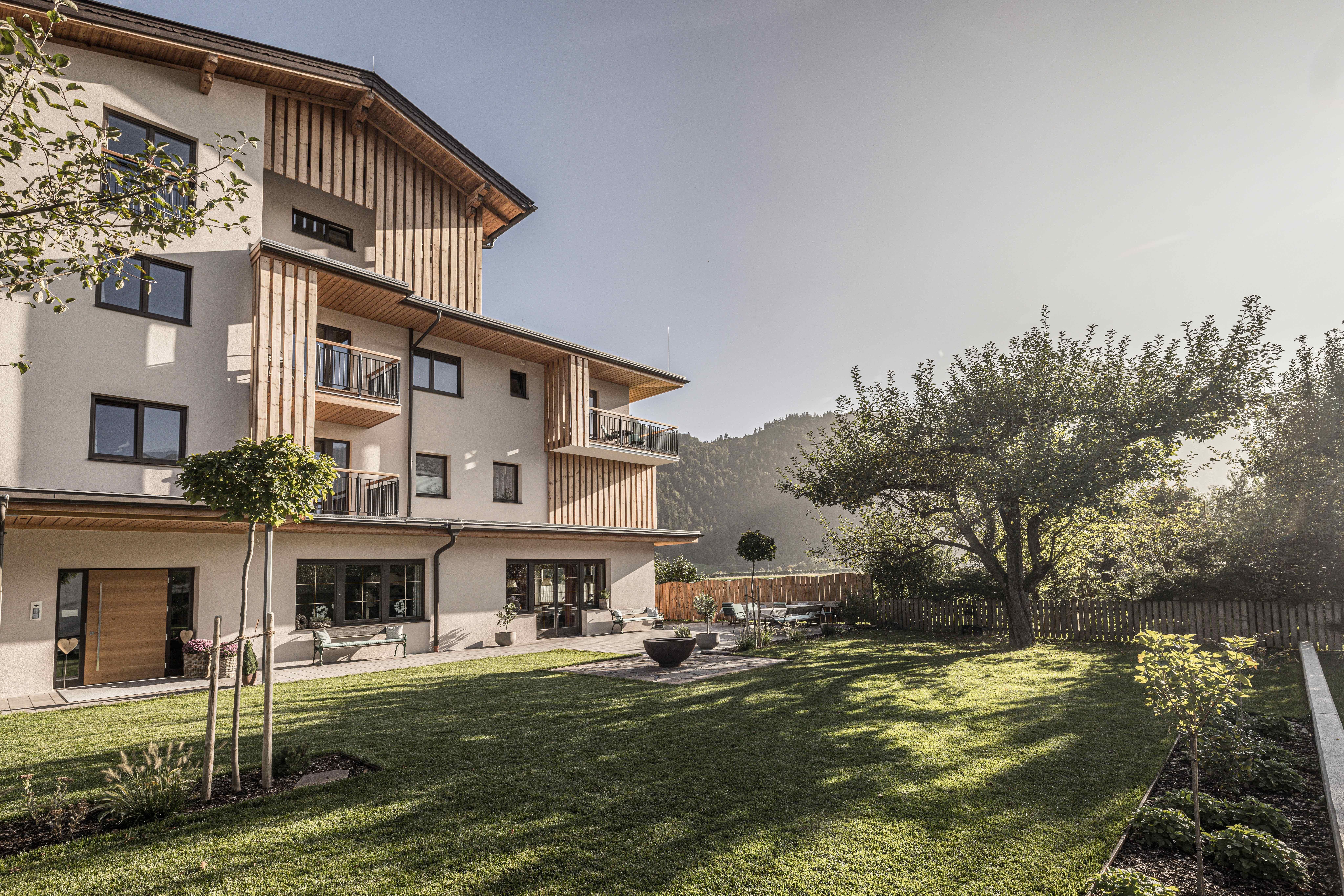 A modern building with wood paneling and balconies. The well-maintained garden is surrounded by trees and offers a beautiful view of the mountains.