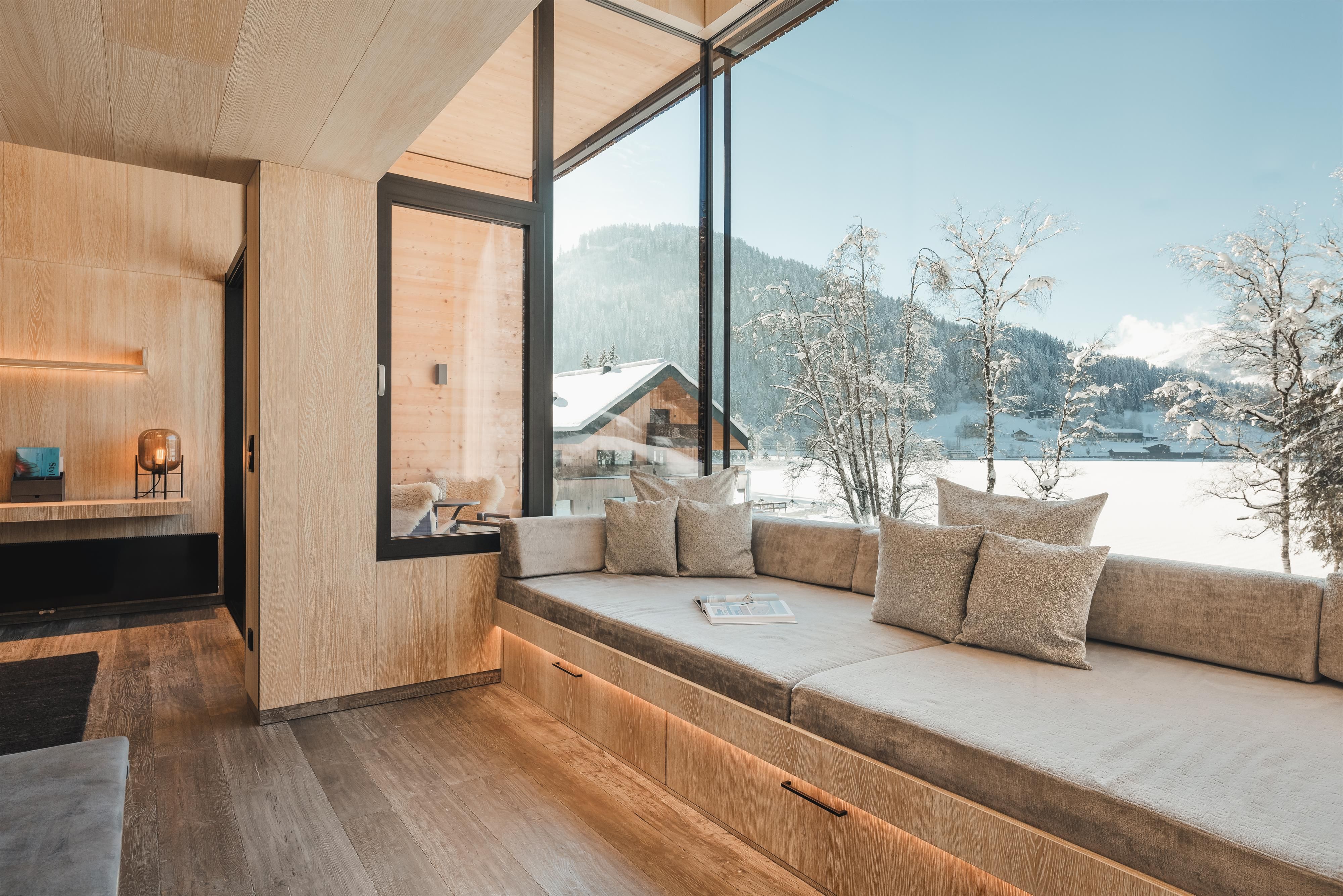 A modern, bright living room with large windows and a view of snowy mountains. The seating area is cozy, designed with cushions and wooden-paneled walls.