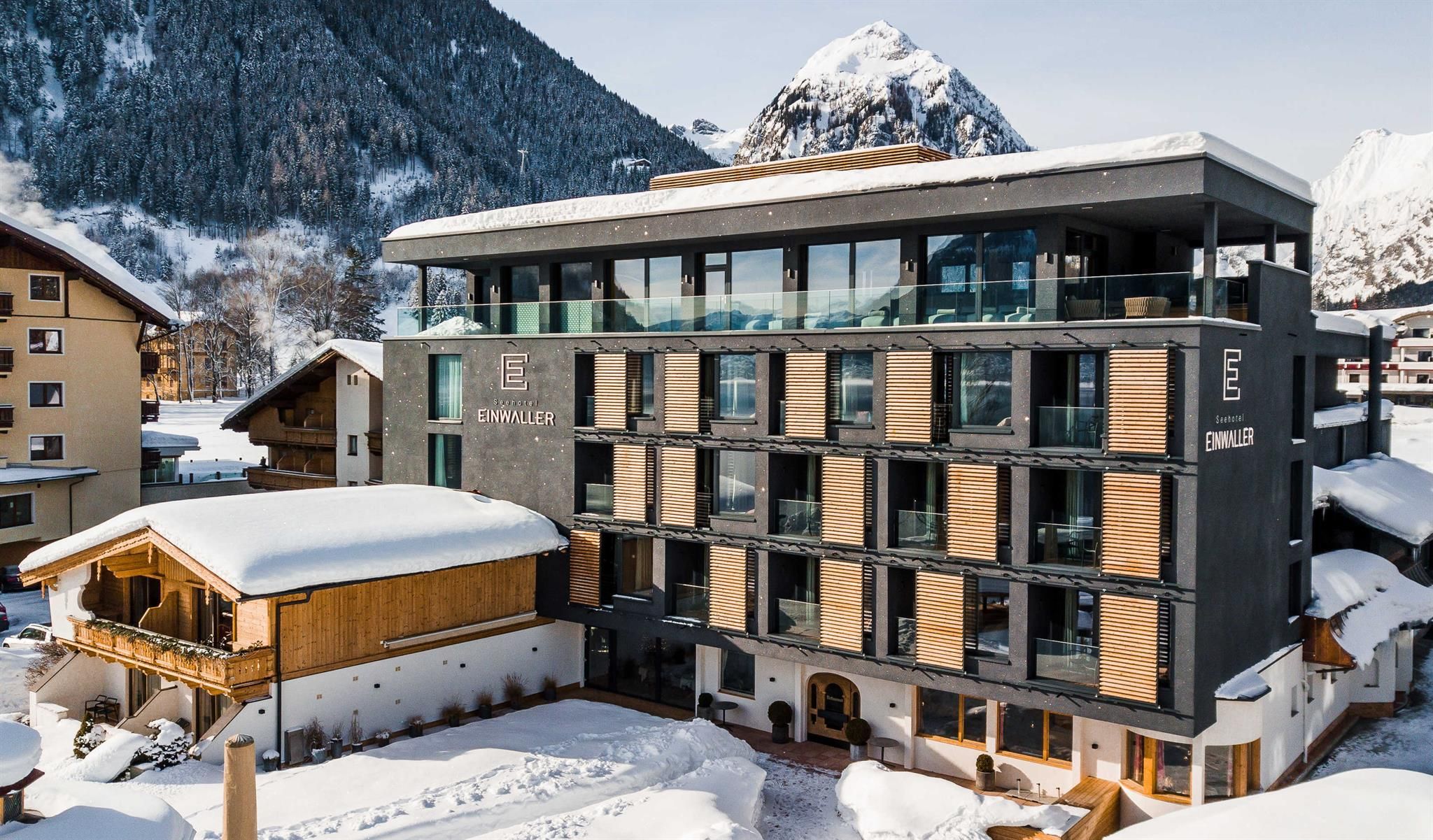 A modern hotel building surrounded by snow-covered mountains. The architecture combines wood and glass and radiates a cozy style.