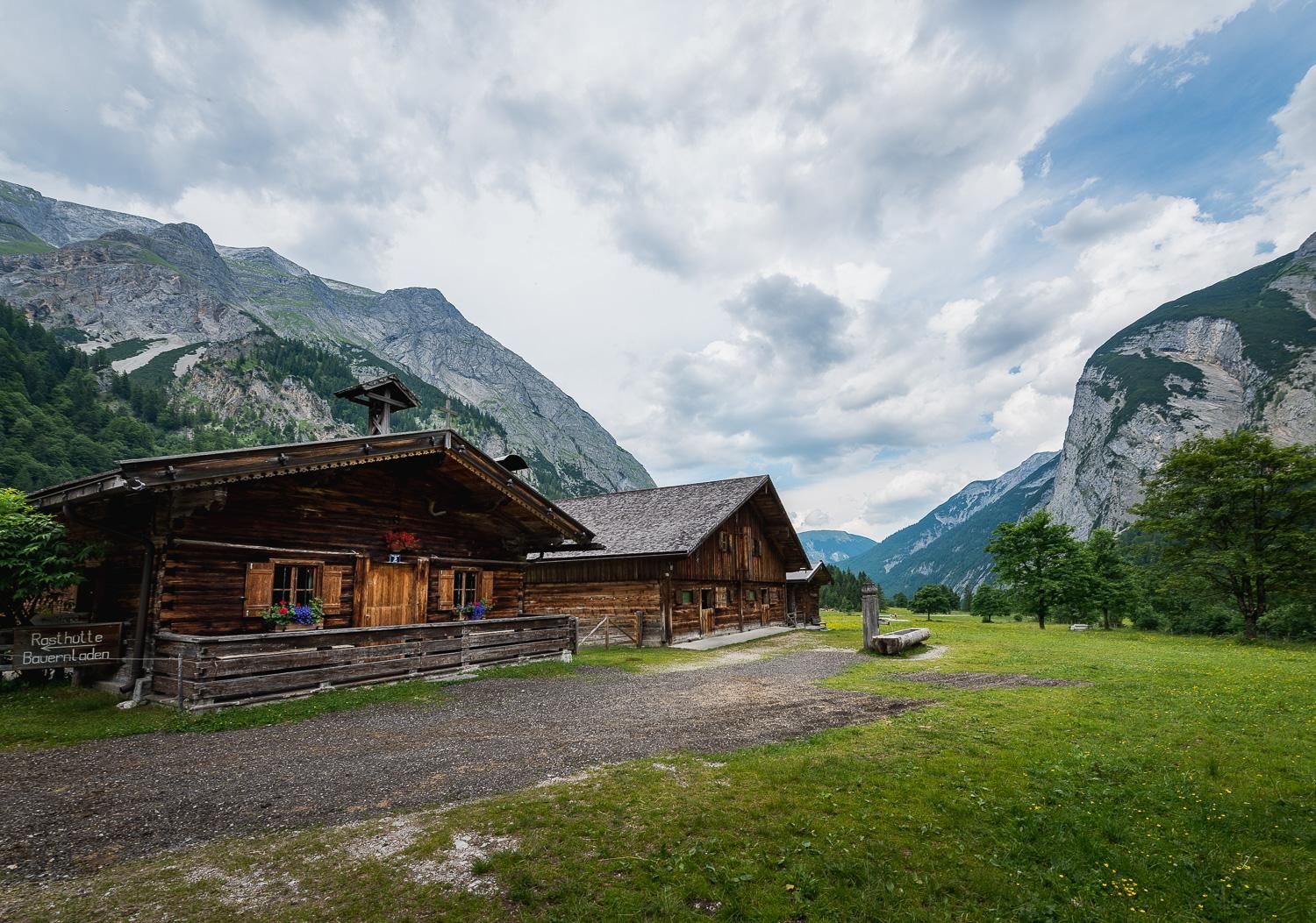 A charming wooden cabin in the midst of the mountains. The surrounding area is green with large rocks and a cloudy sky.