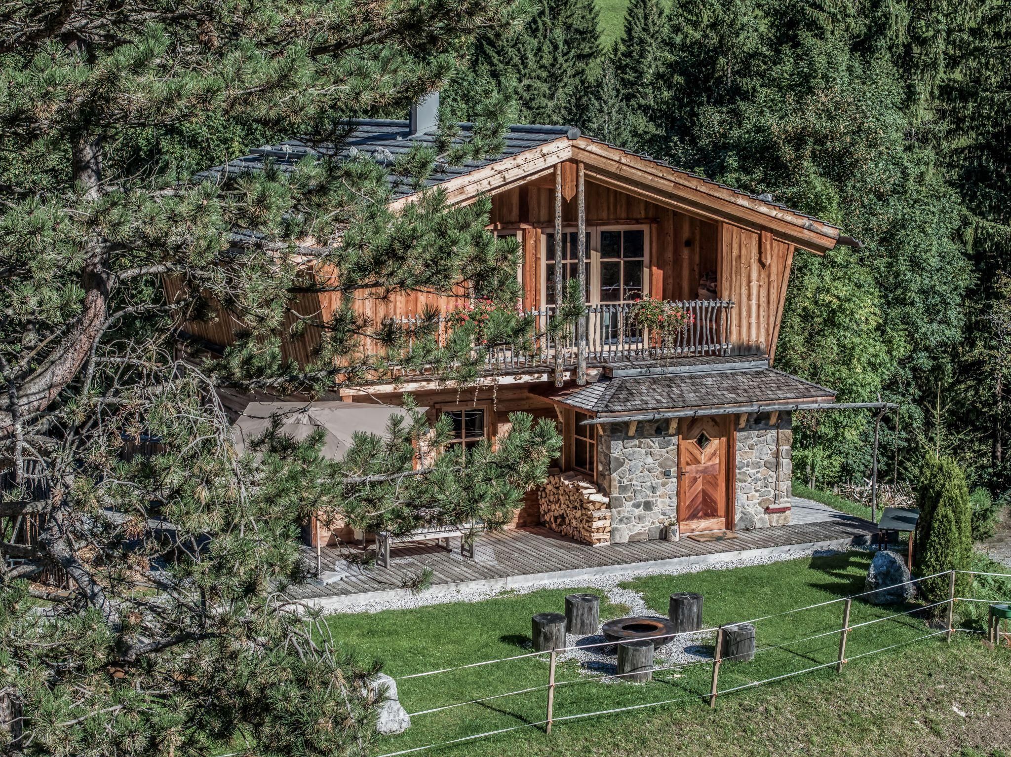 A beautiful chalet surrounded by trees and well-maintained greenery. In front of the house, there is a fire pit with seating options.