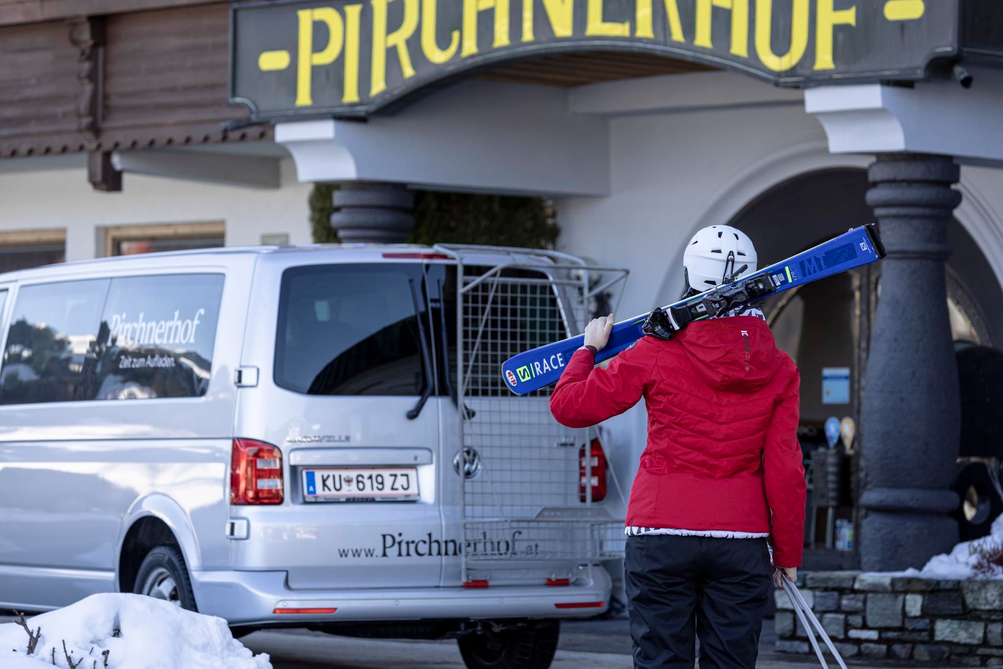 A person with skis is standing in front of a vehicle with the inscription "Pirchnerhof". The image shows a winter landscape with snow-covered ground.