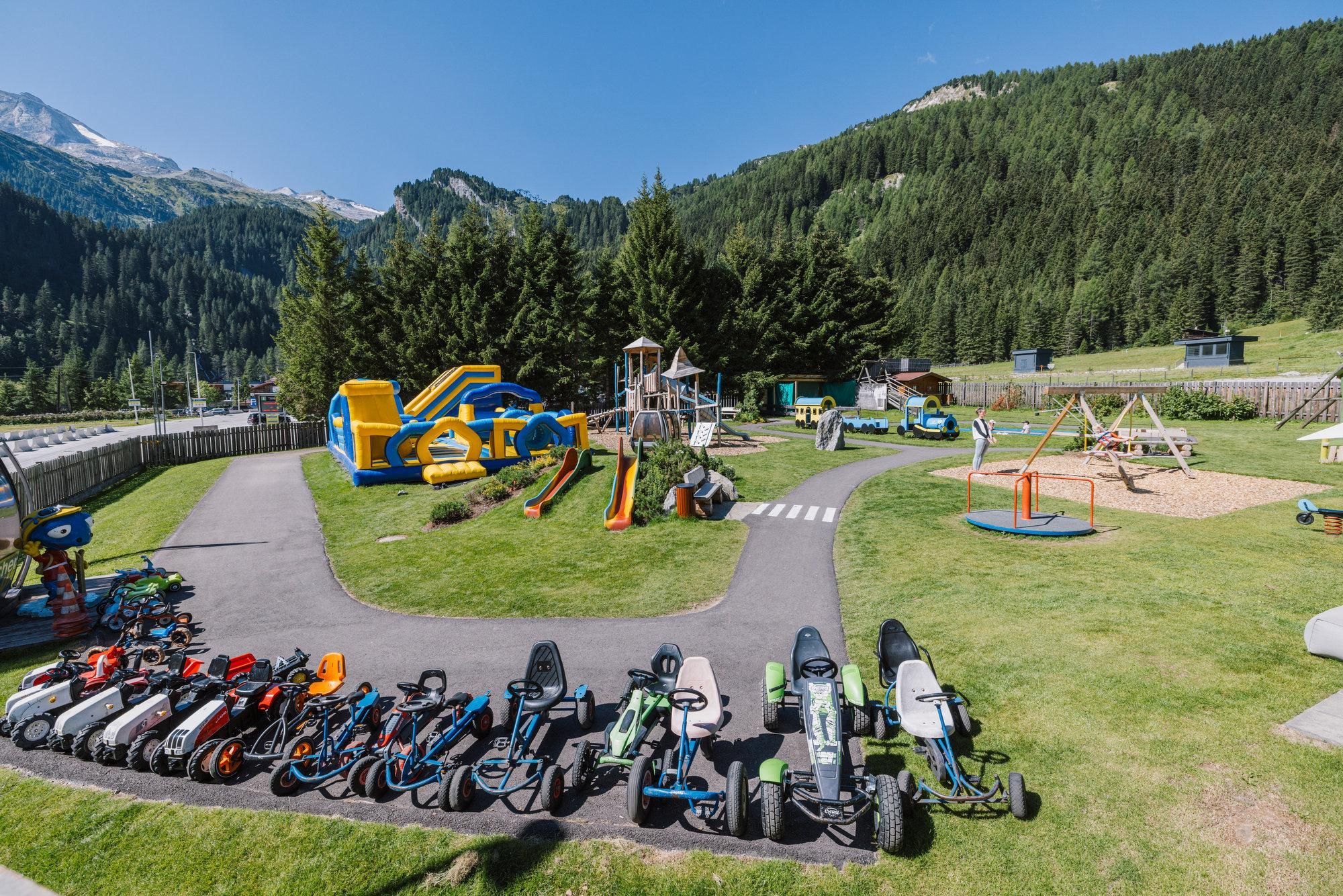A playground with various attractions and an area for children's vehicles. In the background, green mountains and a bright blue sky can be seen.