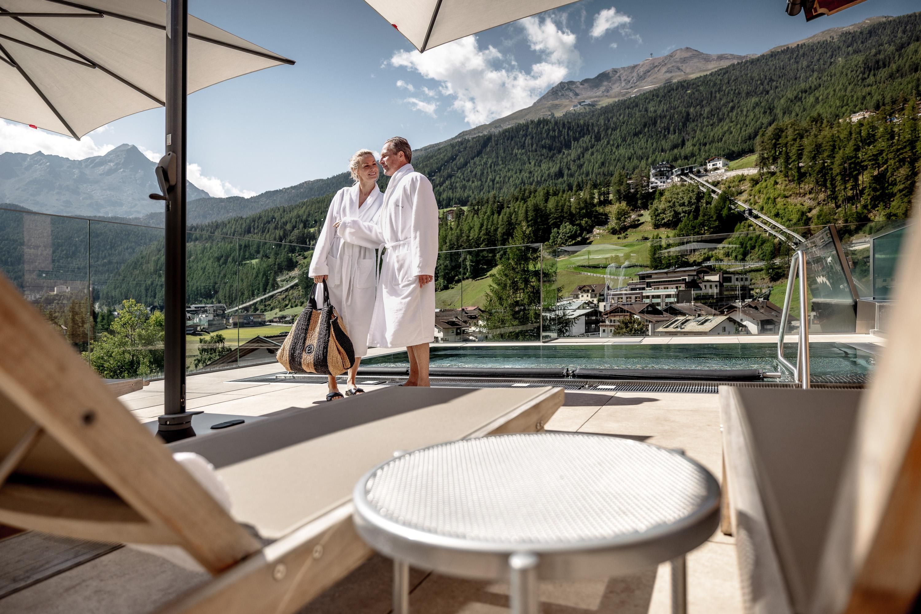 A couple in bathrobes stands by the pool overlooking the mountains. The surroundings are relaxed and luxurious, ideal for a wellness day.