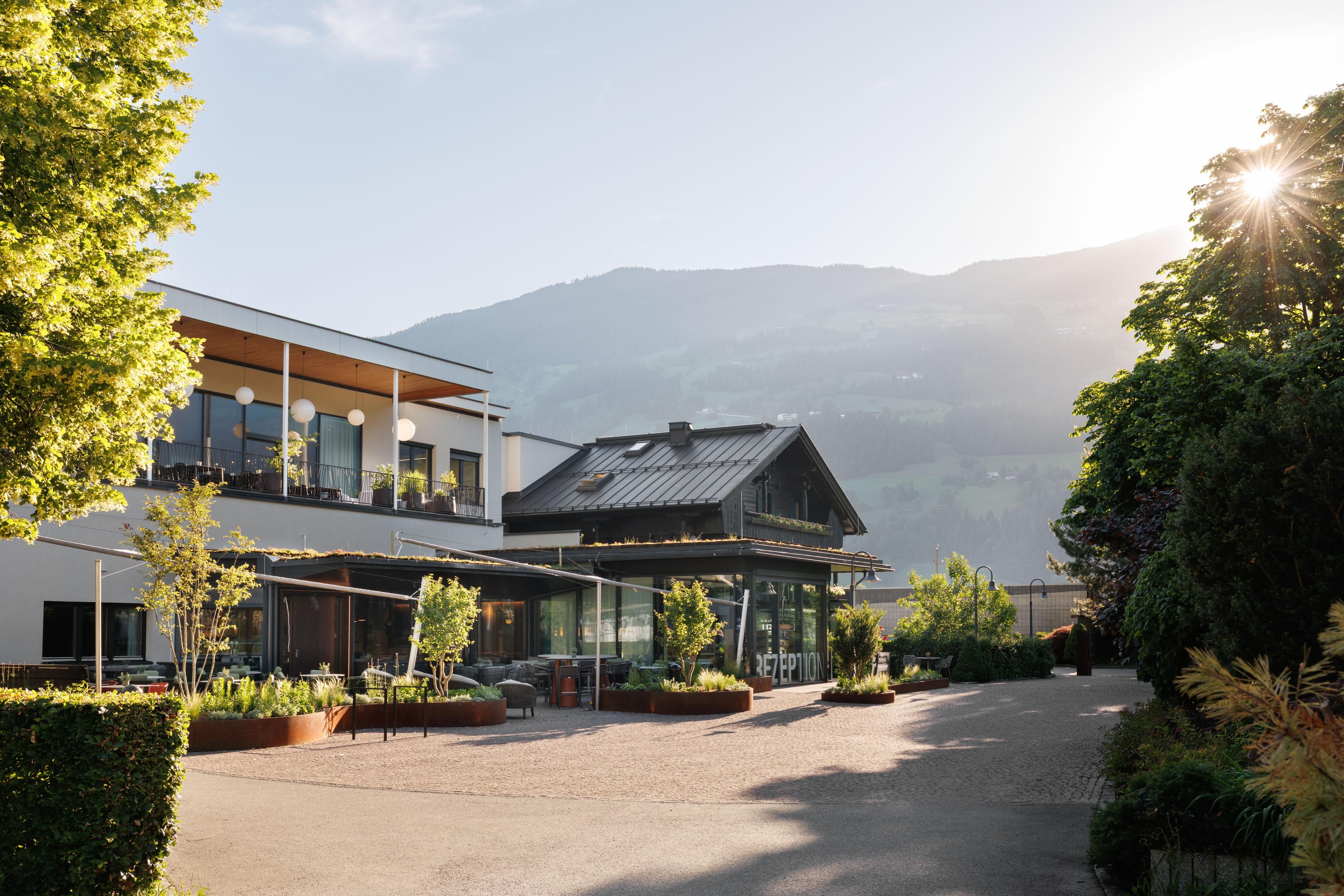 A modern building surrounded by plenty of greenery and mountains in the background. The sun is shining and creating a warm atmosphere.