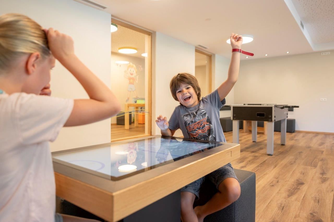 A cheerful child is playing at an interactive table, while another child is distracted. The room has a modern decor with light wooden flooring.