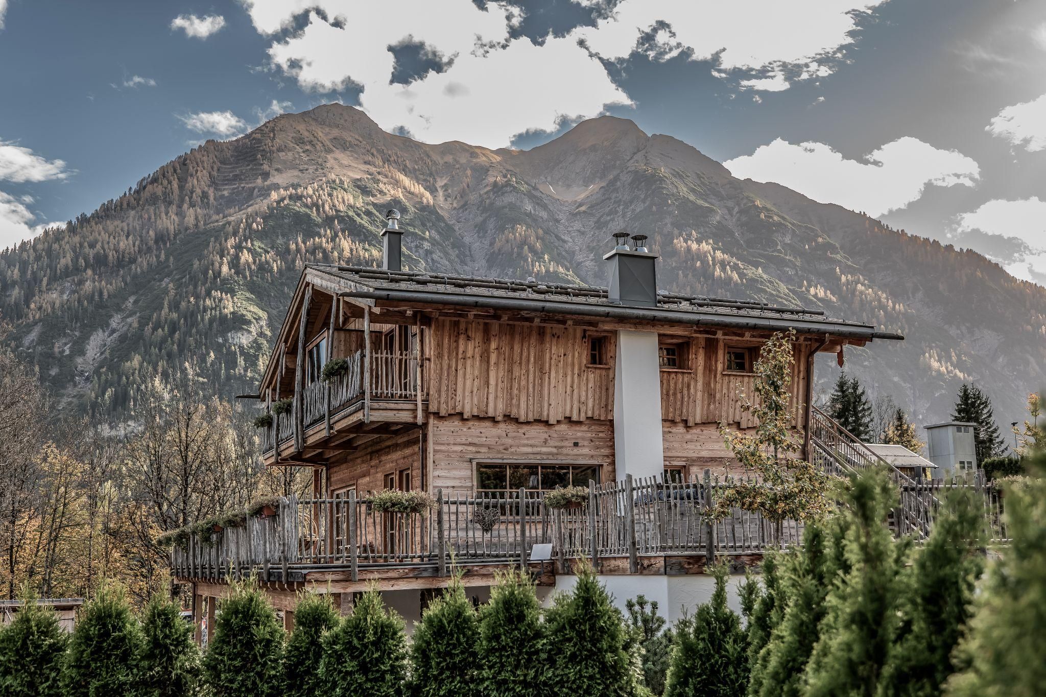 A picturesque chalet with a balcony, surrounded by green trees. In the background, a majestic mountain range rises under a partly cloudy sky.