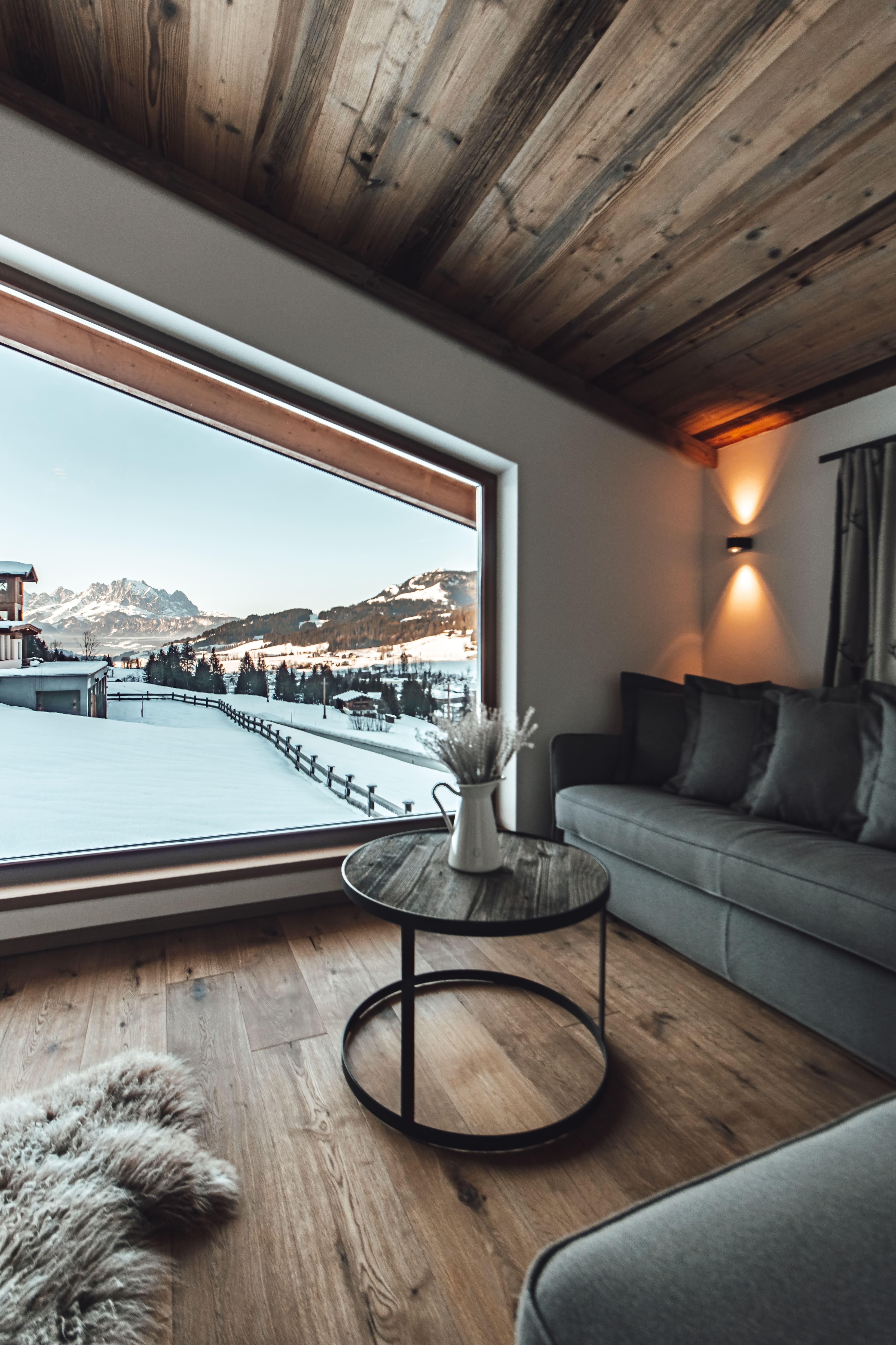 A cozy living space with a large window that shows a snow-covered mountain landscape. The room is modernly furnished with a gray sofa and a round coffee table.