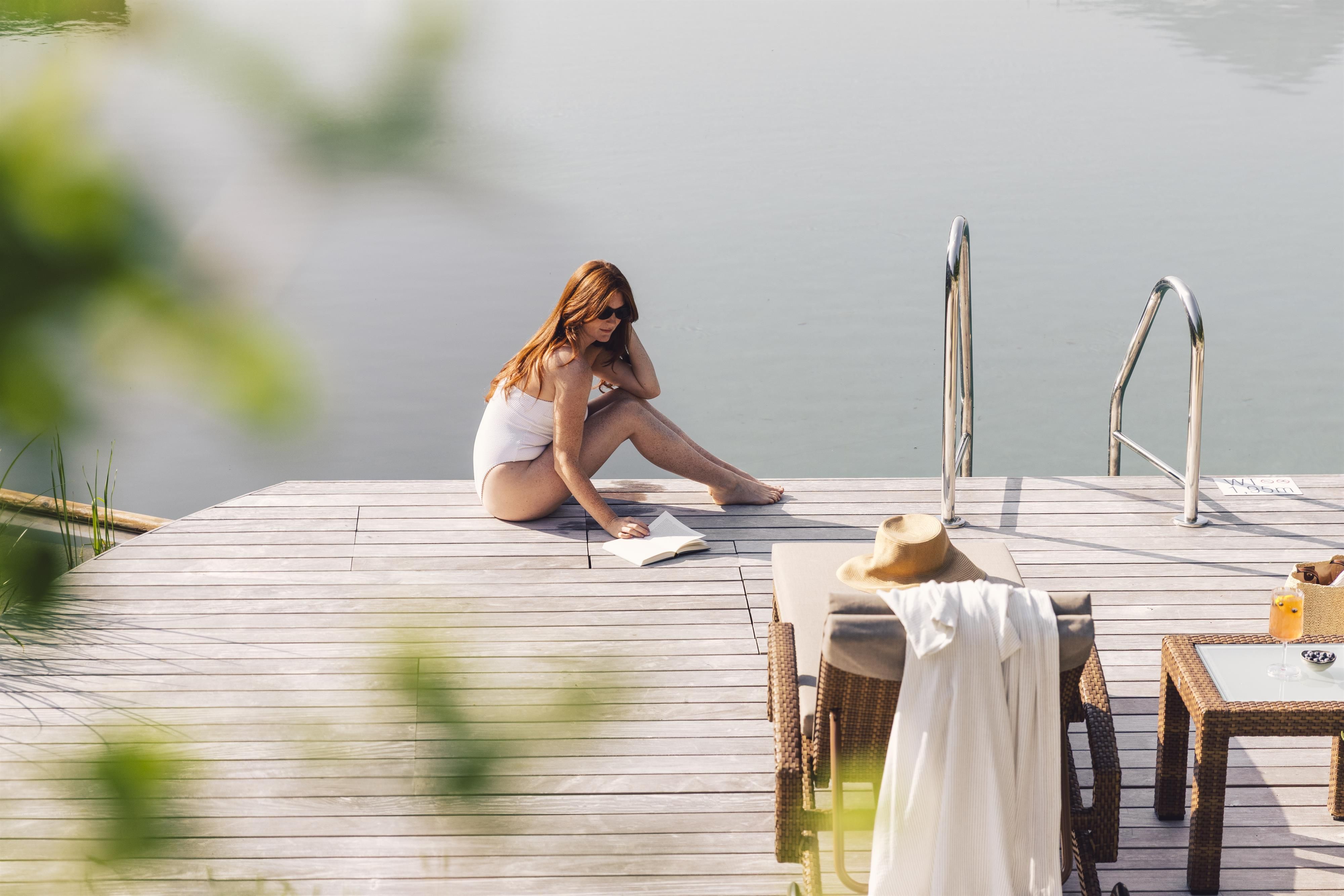 A woman is sitting on a wooden deck by the water, reading a book. Next to her, there is a hat and a towel.
