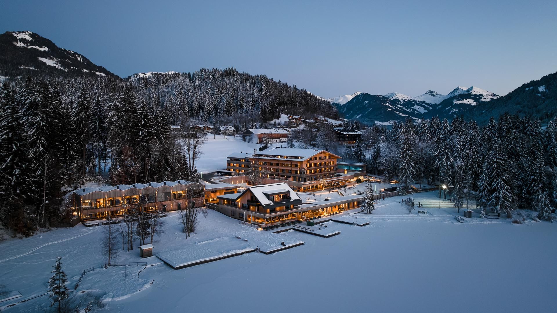 A picturesque winter village, surrounded by snow-covered mountains and fir trees. The buildings are softly illuminated and reflect a cozy atmosphere.