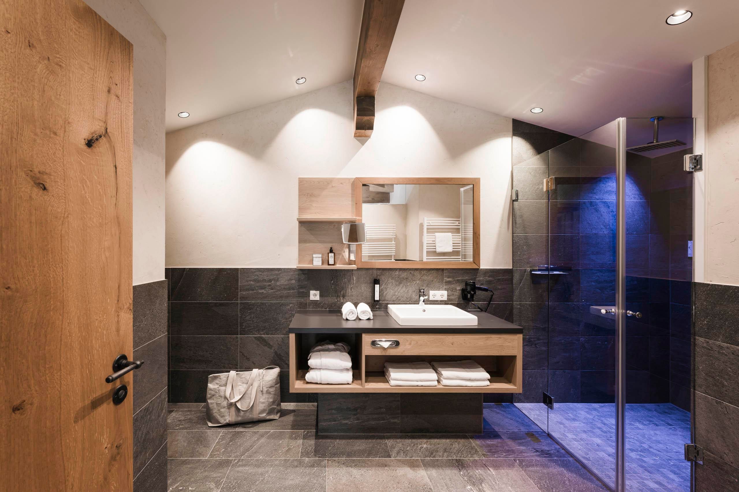 A modern bathroom with a wood paneling and elegant tiles. It features a sink, towels, and a shower.
