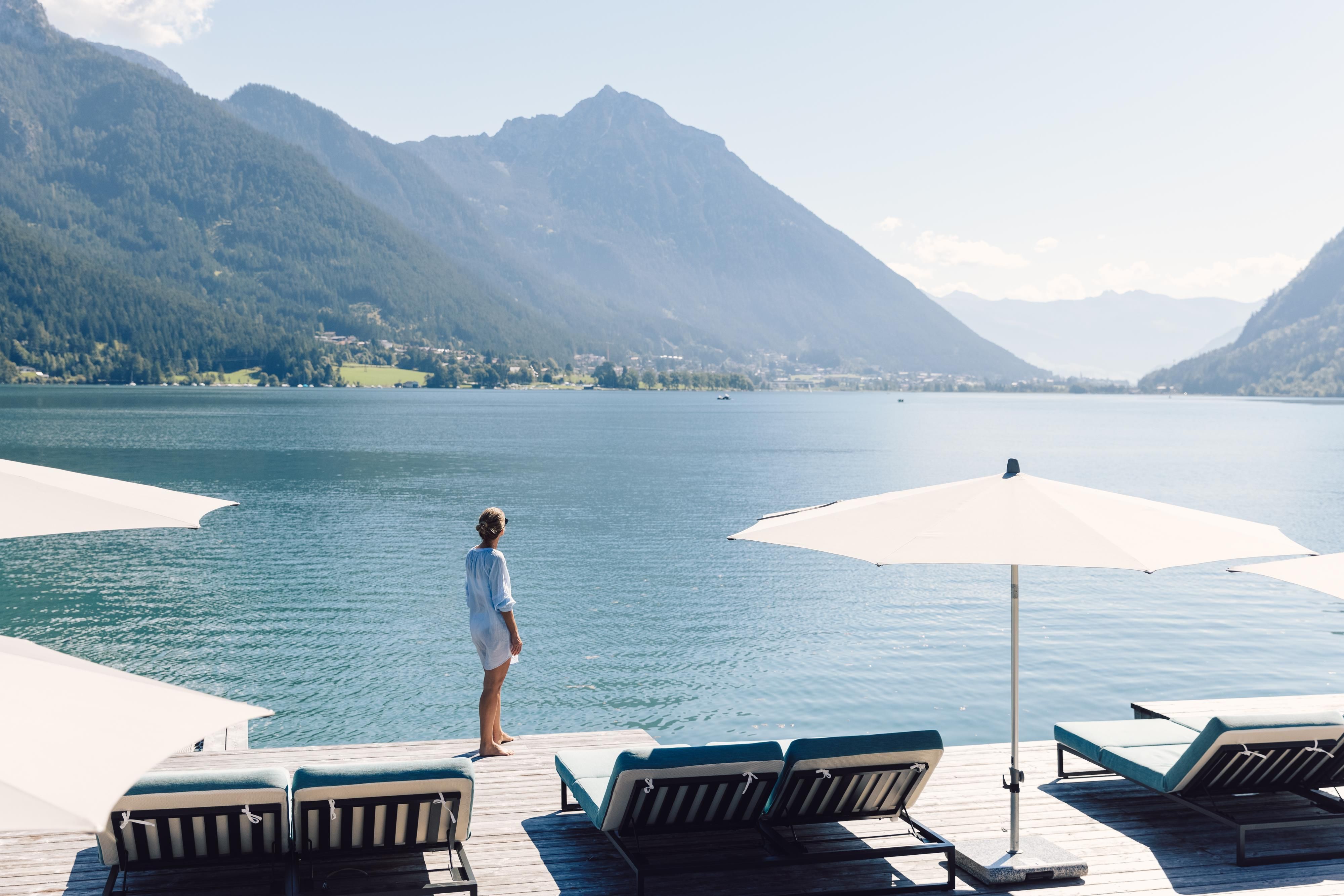 A person stands by the water overlooking the mountains and the lake. Sun loungers and parasols are visible in the foreground.