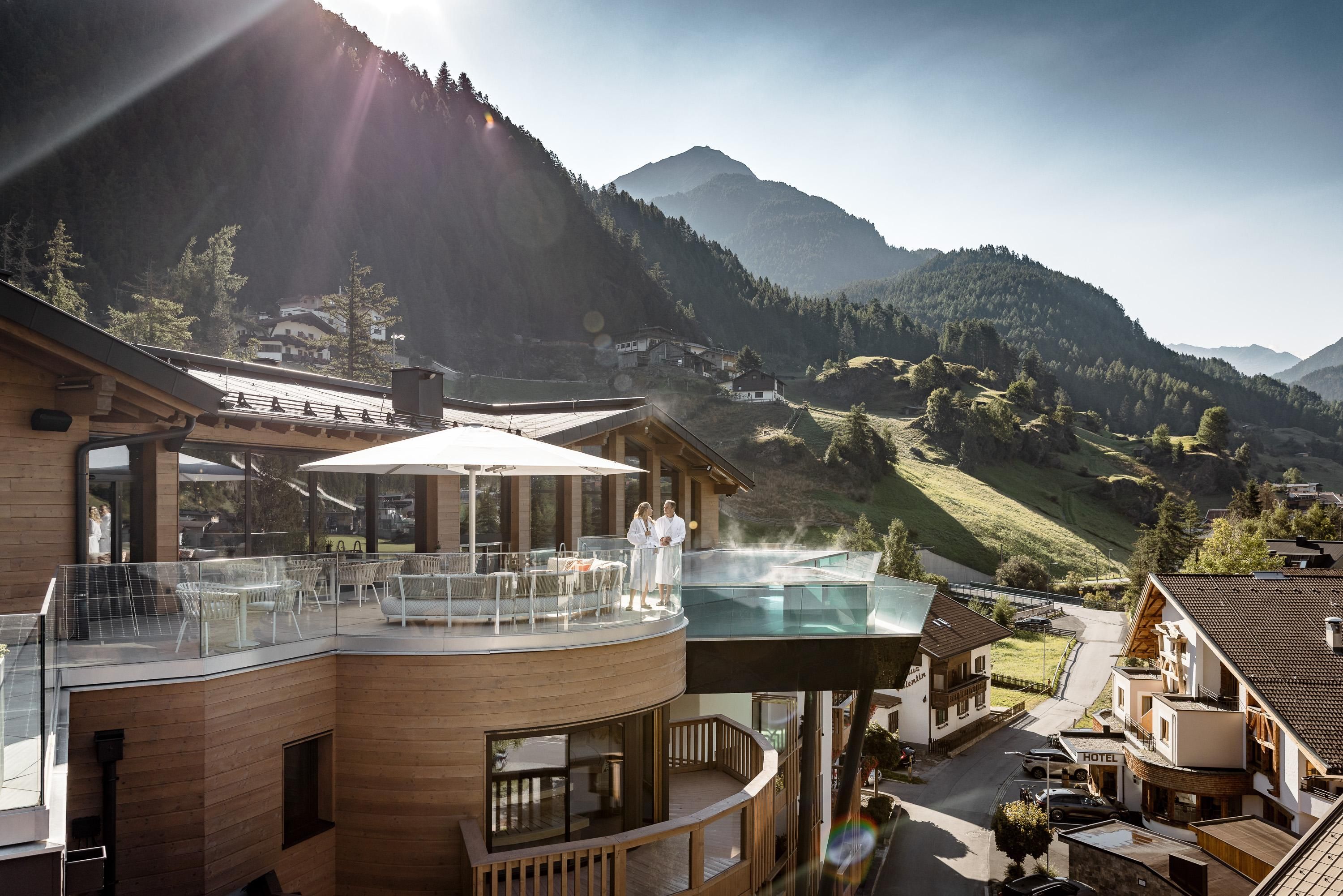 A modern building with a pool on the terrace and a view of the mountains. The surroundings are green and sunny, perfect for a relaxing stay.