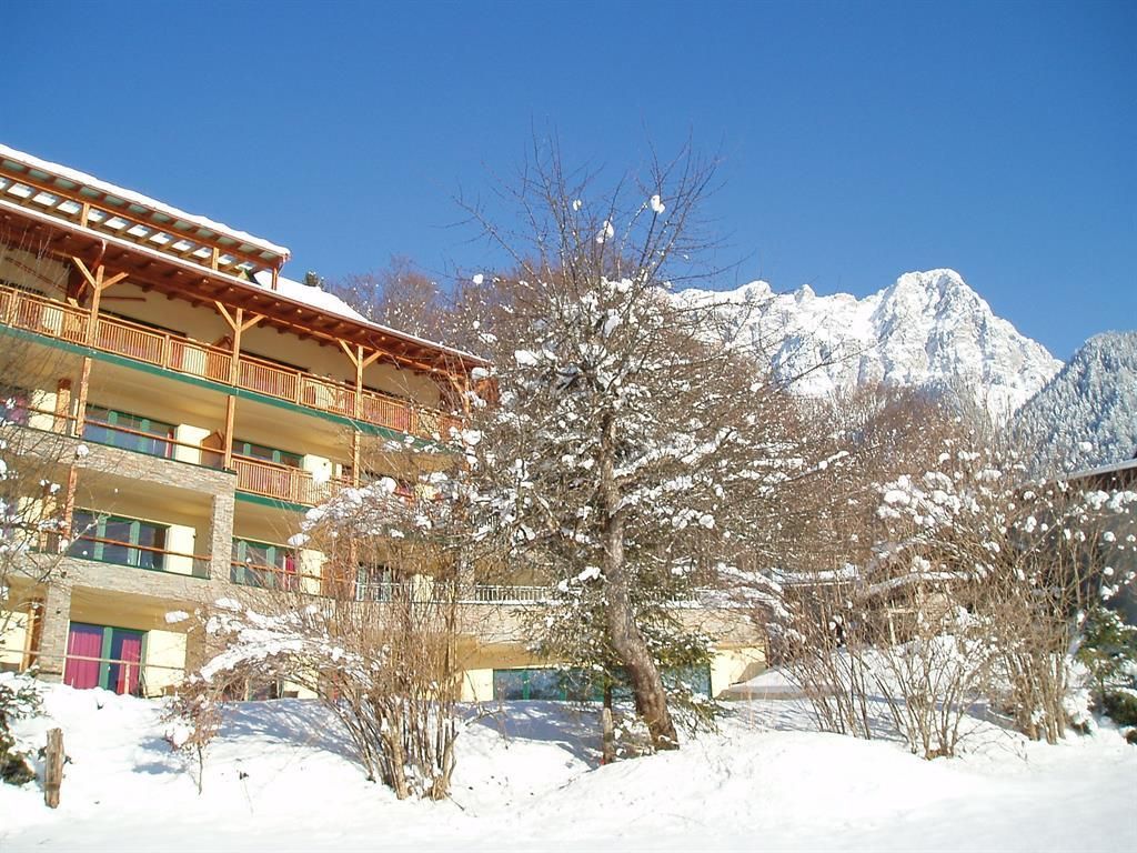 A beautiful winter scene with a modern building, surrounded by snow-covered trees. In the background, impressive mountains can be seen under a clear blue sky.