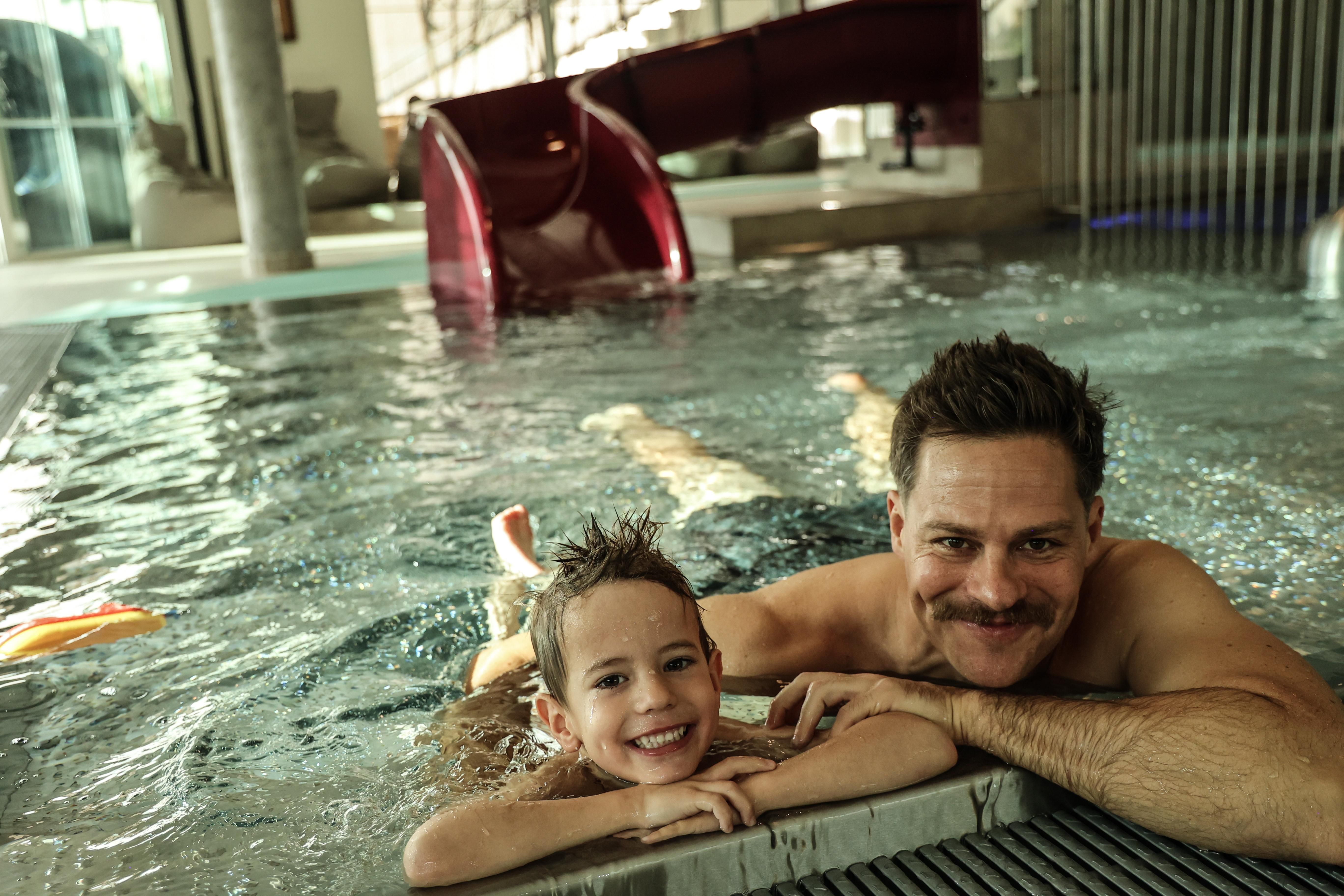 A father and his son enjoy swimming in an indoor pool. Water slides are visible in the background.