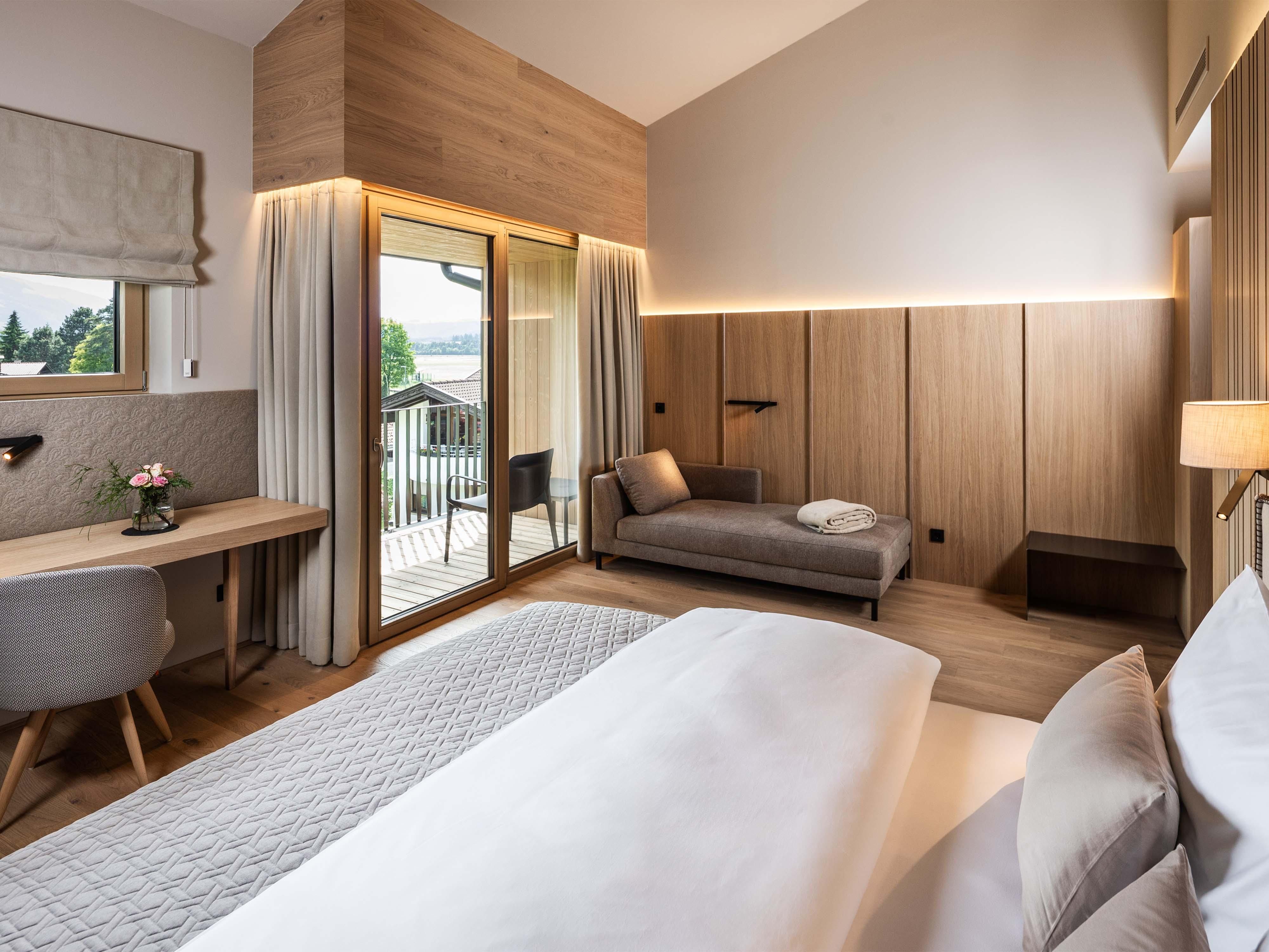 A modern bedroom with wood paneling and large windows. The room is bright and cozy furnished, with a workspace and a sofa.