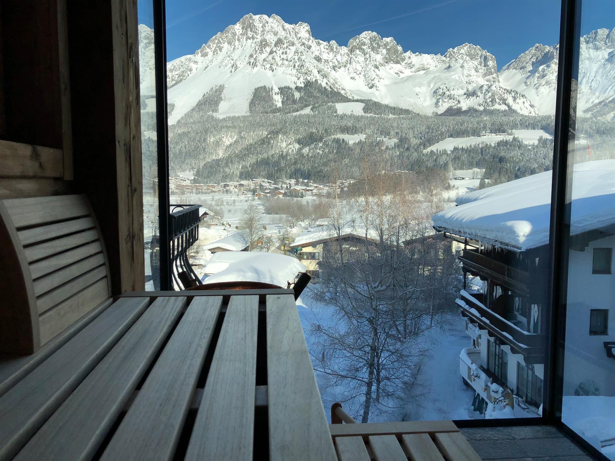 A view from a window of impressive snow-covered mountains. In the foreground, there is a wooden bench that conveys a cozy atmosphere.