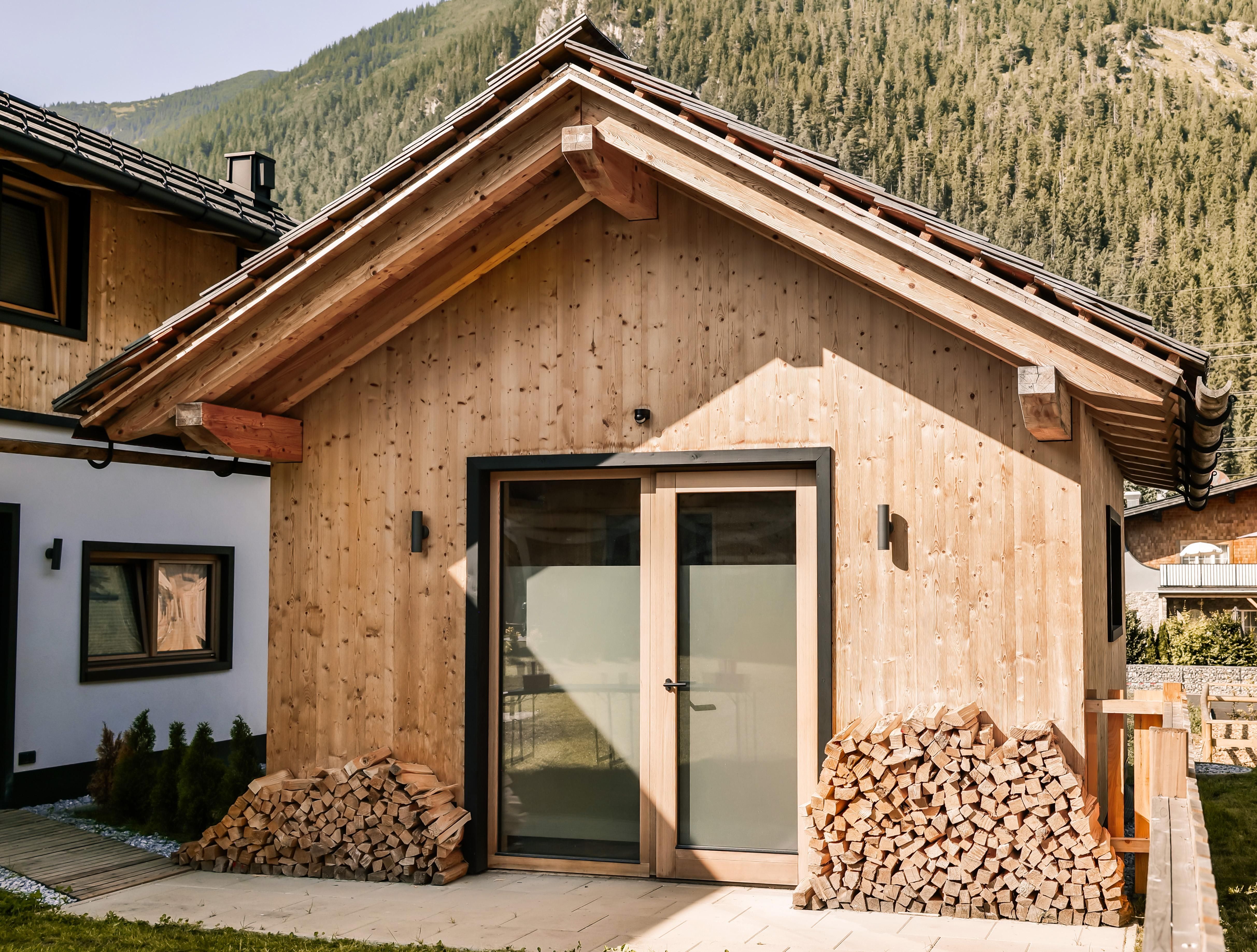 A modern wooden house with a large glass door. In front of the house is a pile of firewood and the surrounding landscape is characterized by mountains.
