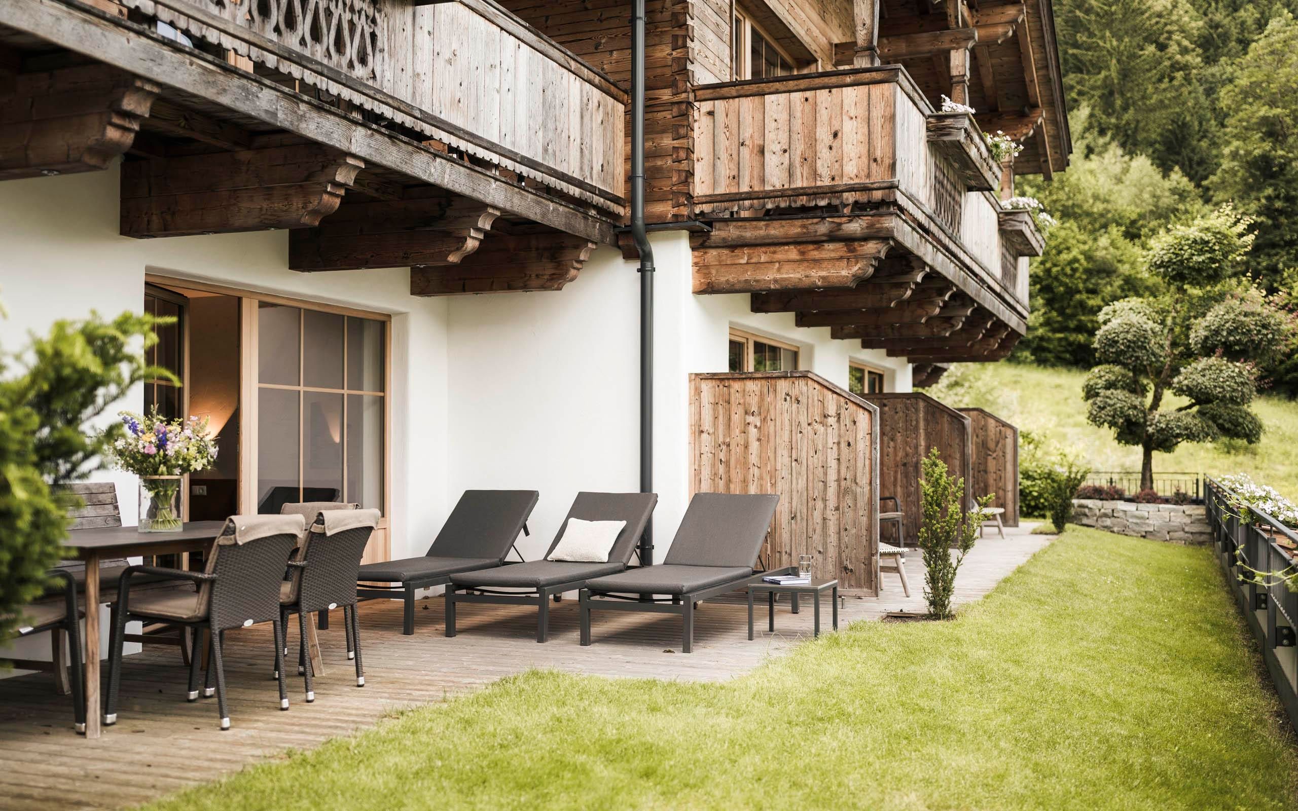 A cozy terrace with loungers and a dining table, surrounded by green meadows. In the background, there are wooden balconies and a beautiful view of nature.