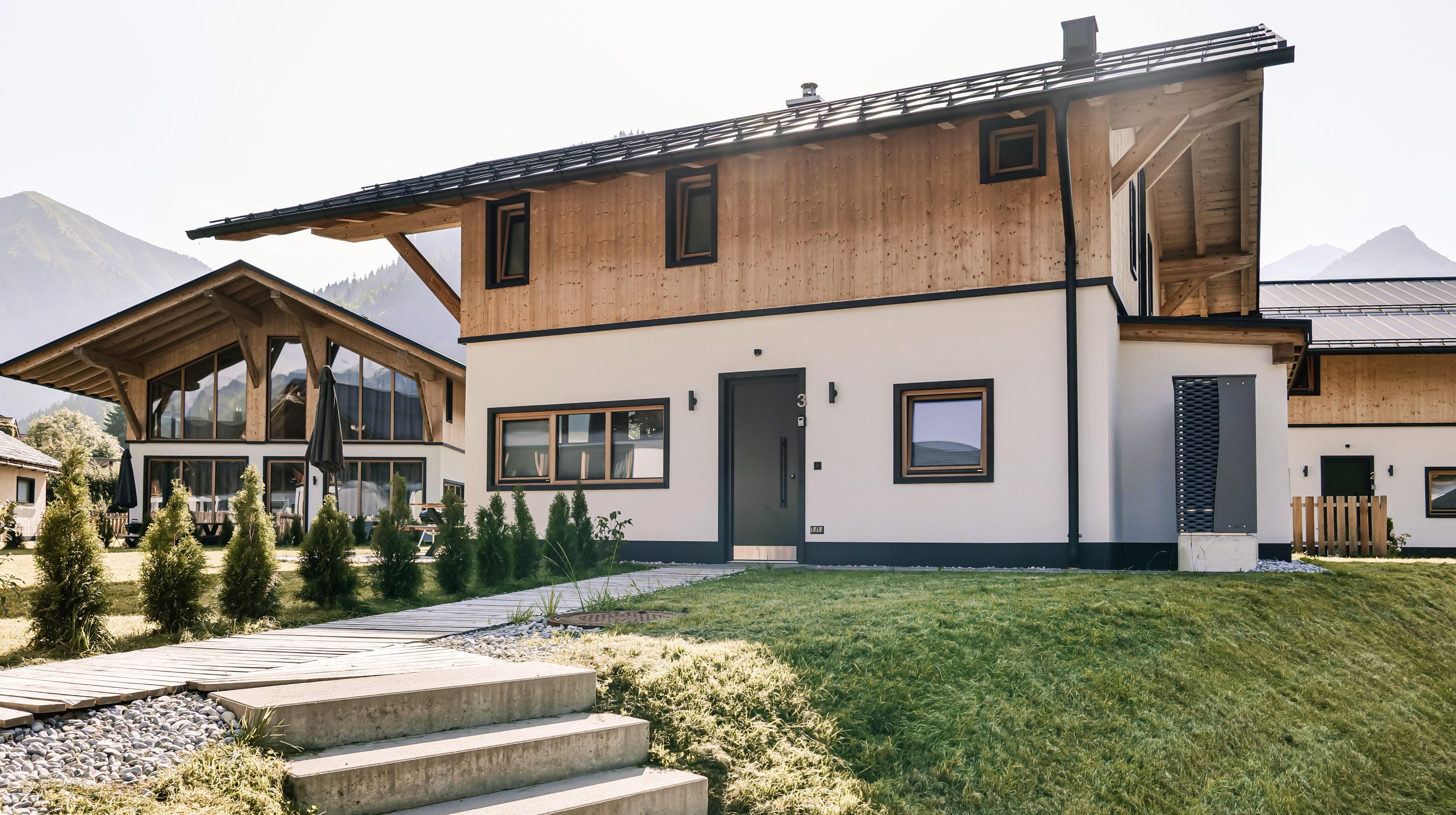 A modern house with a wooden facade and simple windows. In the background, more buildings and mountains can be seen.