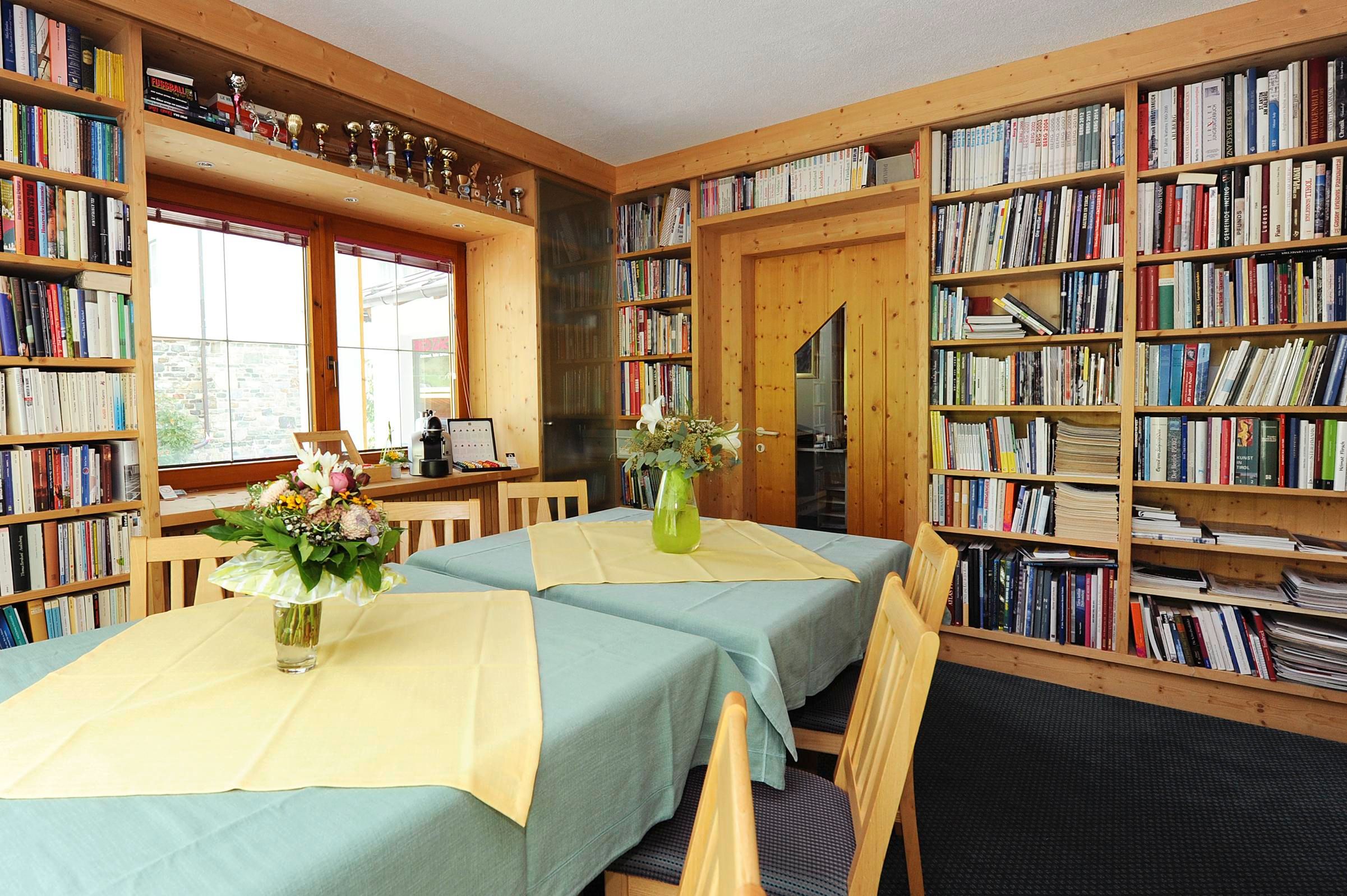 A cozy room with several tables and bookshelves on the walls. A vase of flowers is on one table and the windows let in a lot of light.