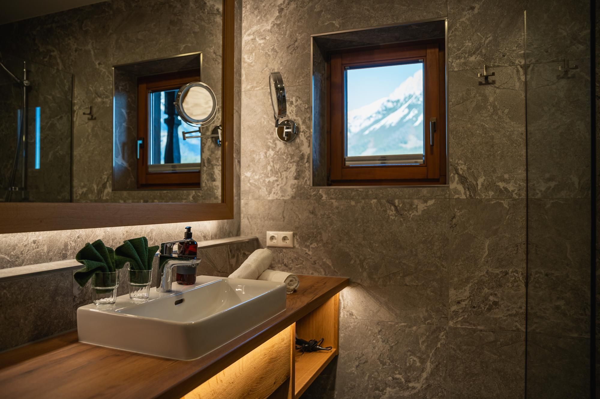 A modern bathroom with an elegant sink and wooden shelves. An impressive mountain landscape can be seen through the window.