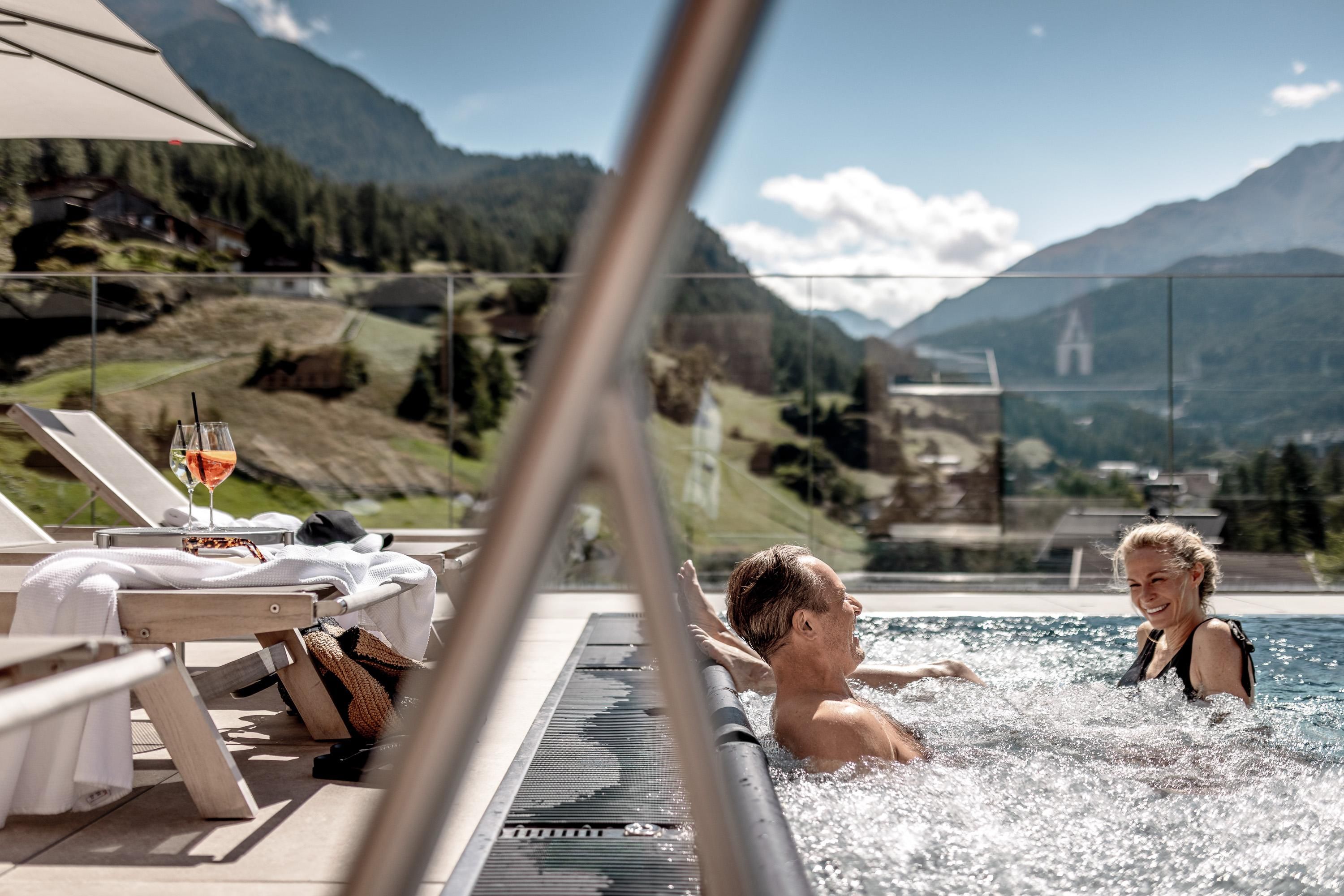 A couple relaxes in the hot tub with a view of the mountains. Next to the pool are loungers and a drink.