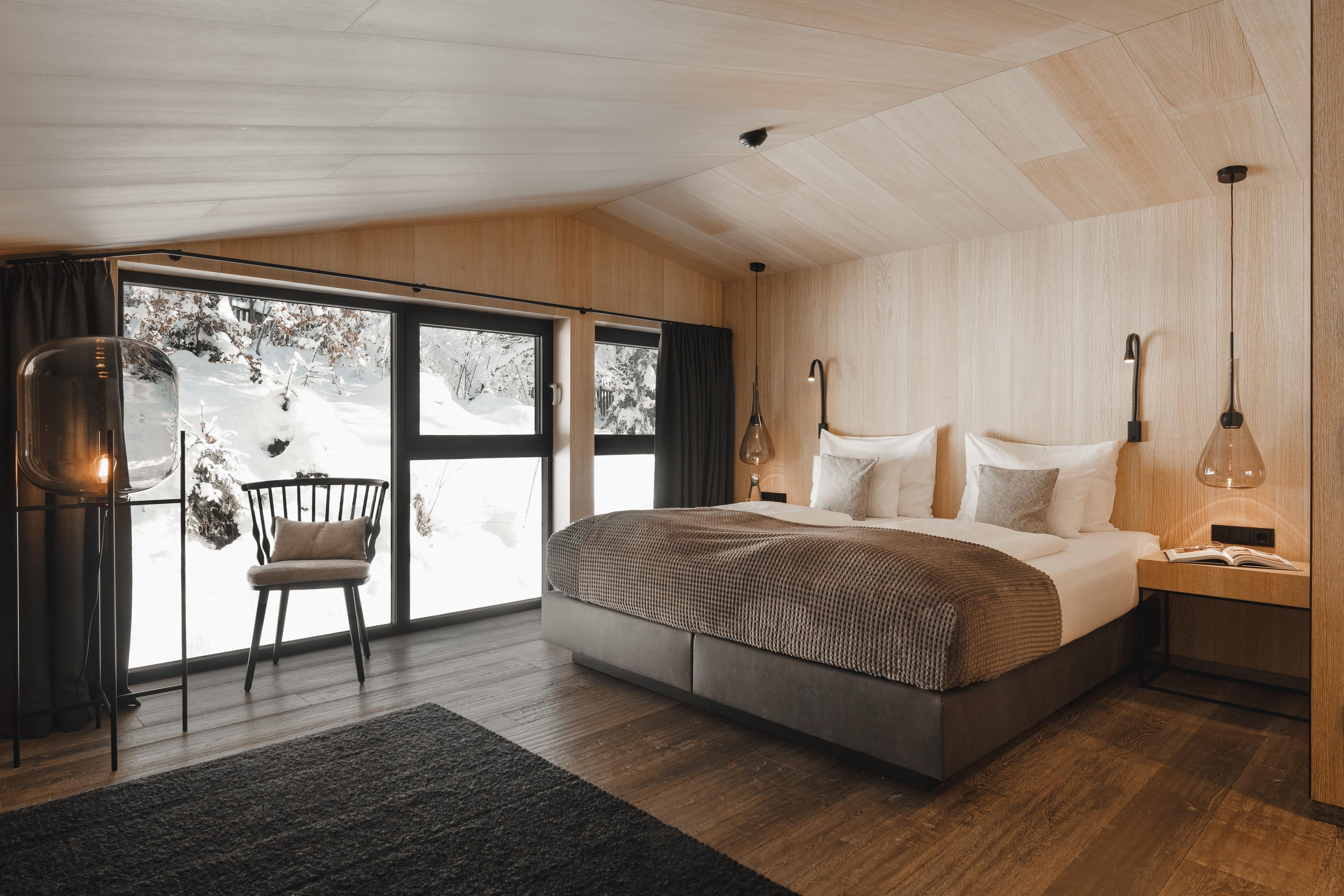 A modern bedroom with wood paneling and large windows. The view shows snowy landscapes and a cozy interior.