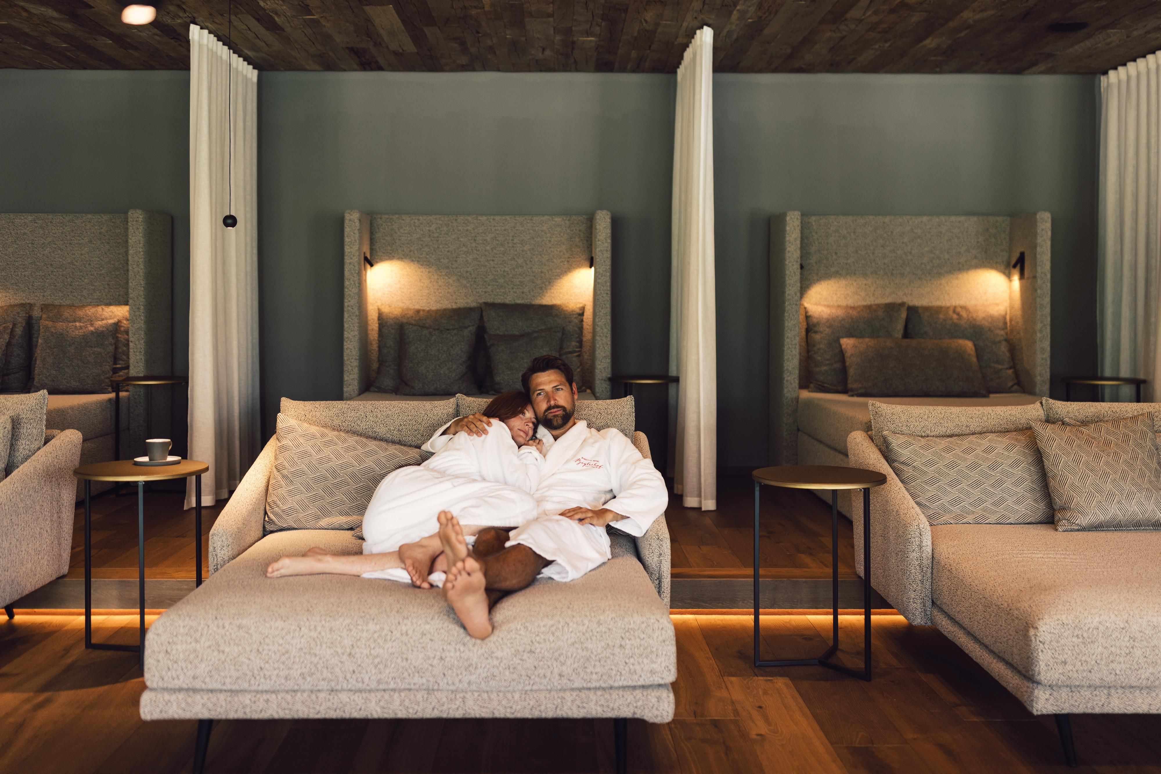 A relaxed man in a bathrobe is lying on a comfortable couch in a modern room. In the background, more cozy loungers can be seen.