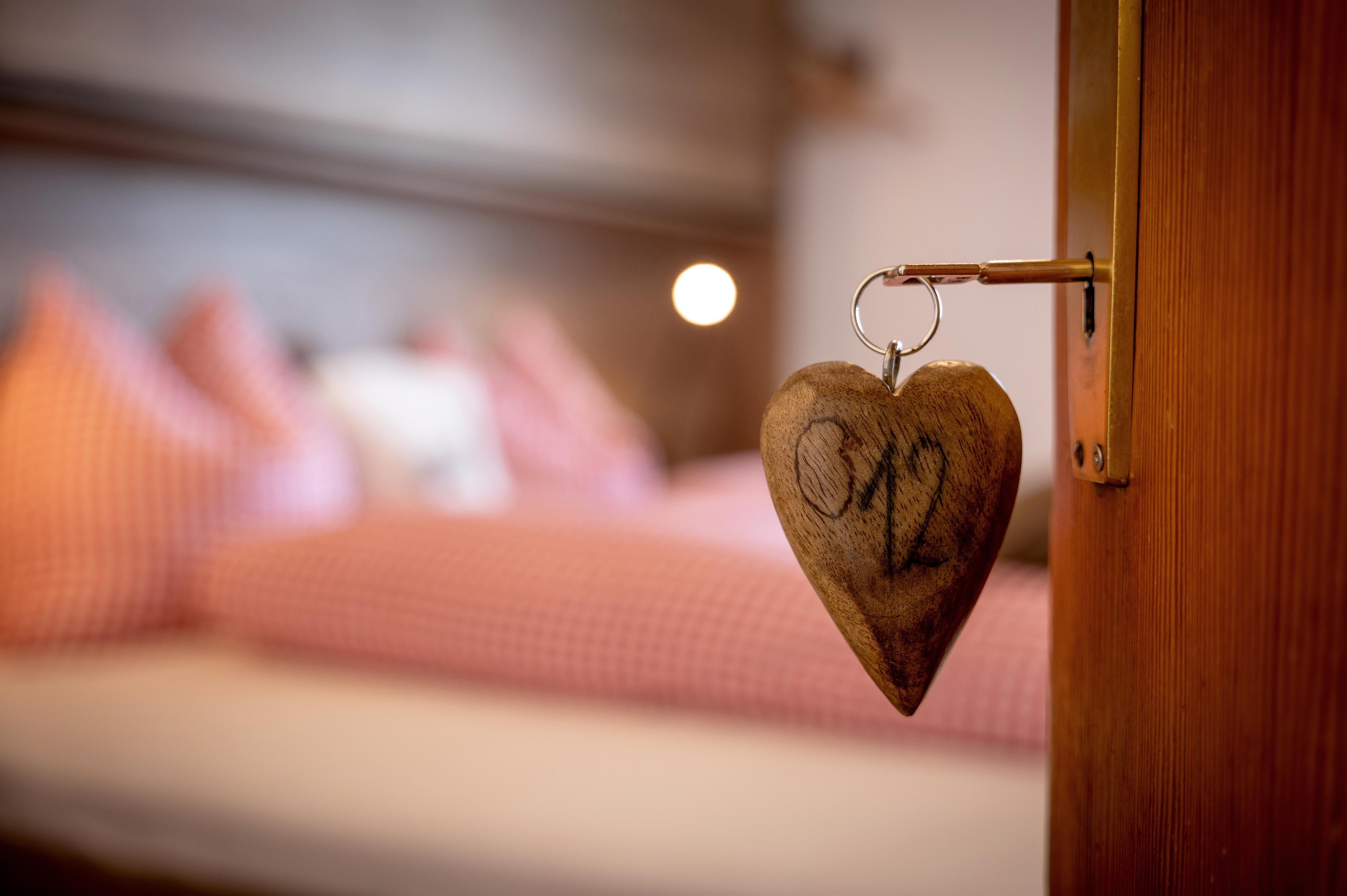 A wooden heart with the number "02" hangs on a room door. In the background, a cozy bedroom with checkered pillows can be seen.