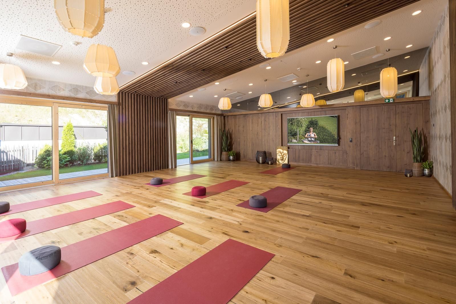 A beautiful, bright yoga room with wooden floors and red mats. Large windows bring in plenty of light and offer a view of the outside.