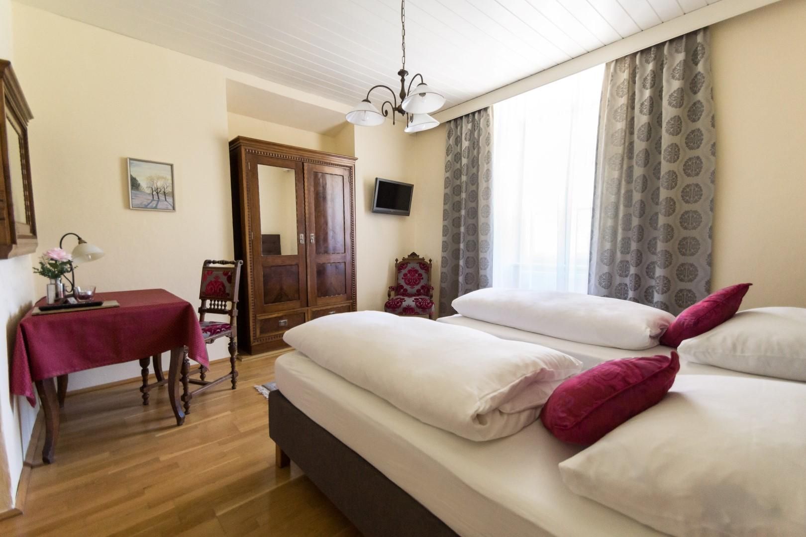 A cozy bedroom with a comfortable double bed and soft colors. The room is furnished with a table, a chair, and a large window.