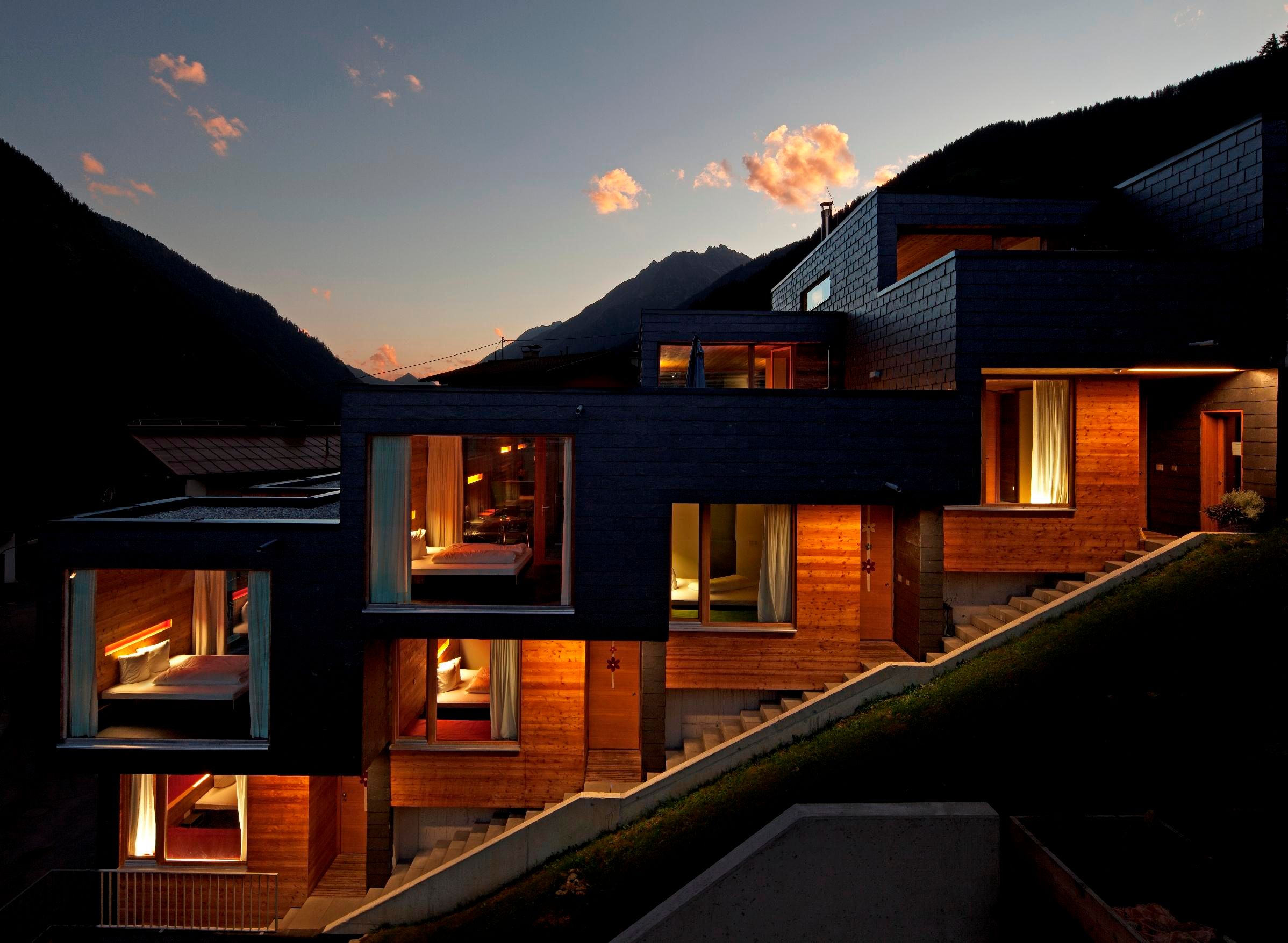 A modern building with a wooden facade at twilight. The windows emit warm light and are surrounded by mountains.
