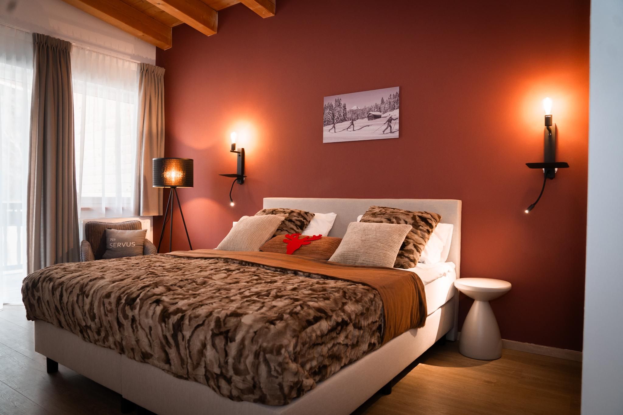 A cozy bedroom with a large bed and soft pillows. The walls are red, and there is nice lighting as well as a view outside.
