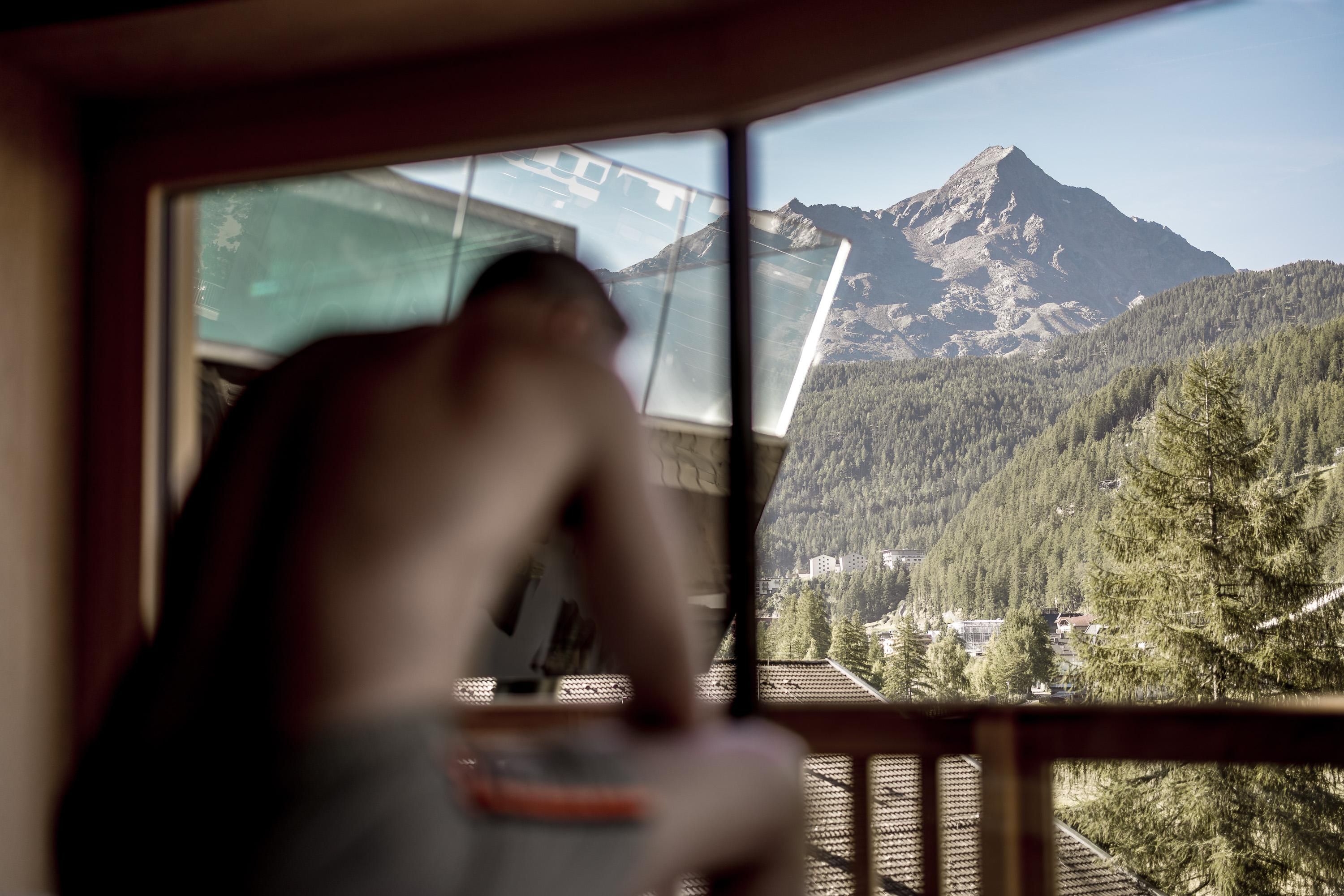A man sits by the window overlooking an impressive mountain landscape. The nature around him is green and serene.