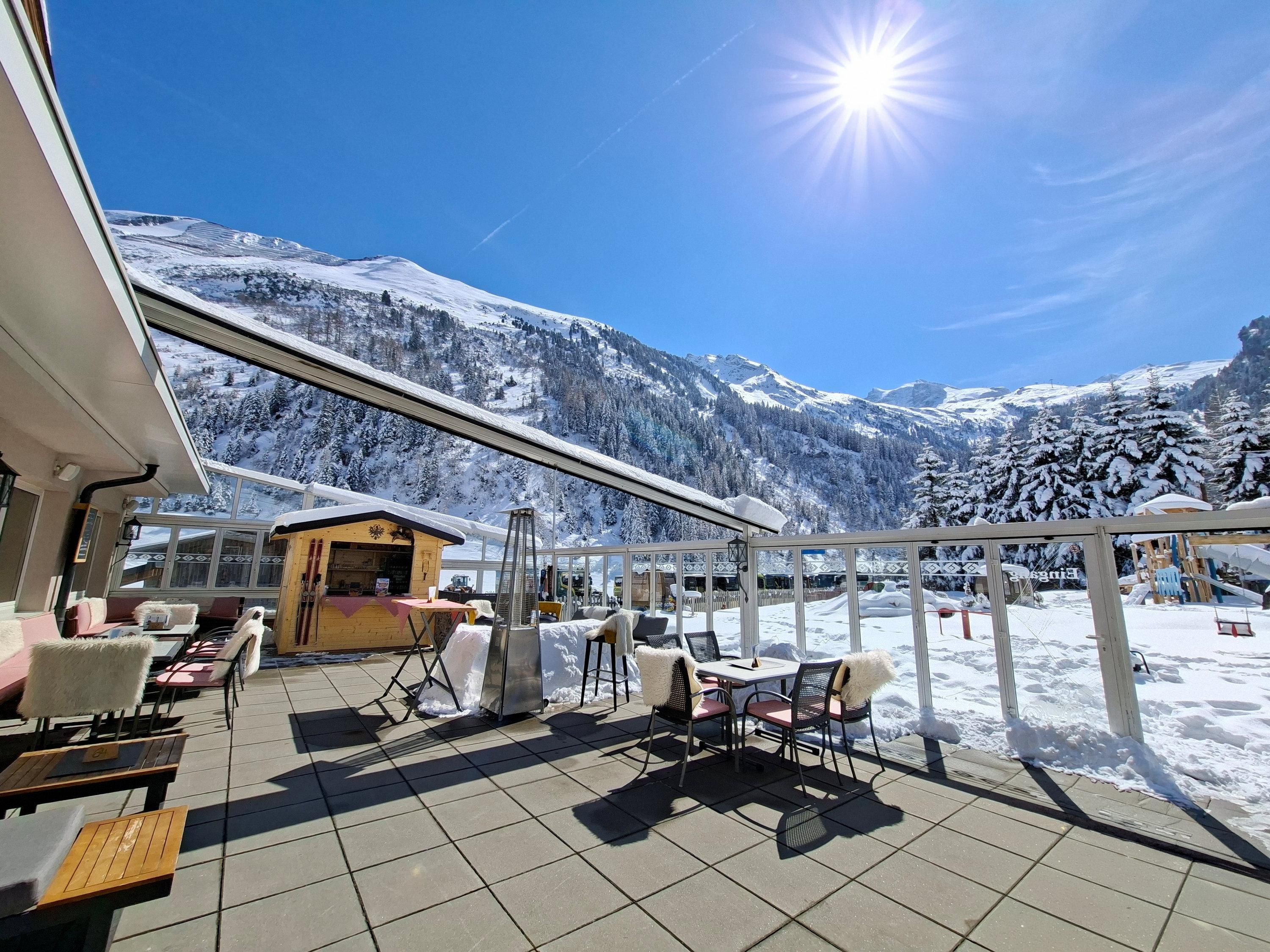 A sunny terrace with a view of snow-covered mountains. The sky is clear and blue, perfect for a relaxing stay outdoors.